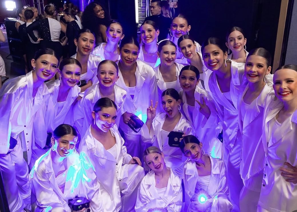 Group of young girls in white costumes smiling, some holding glowing objects, in a dark setting with purple lighting.