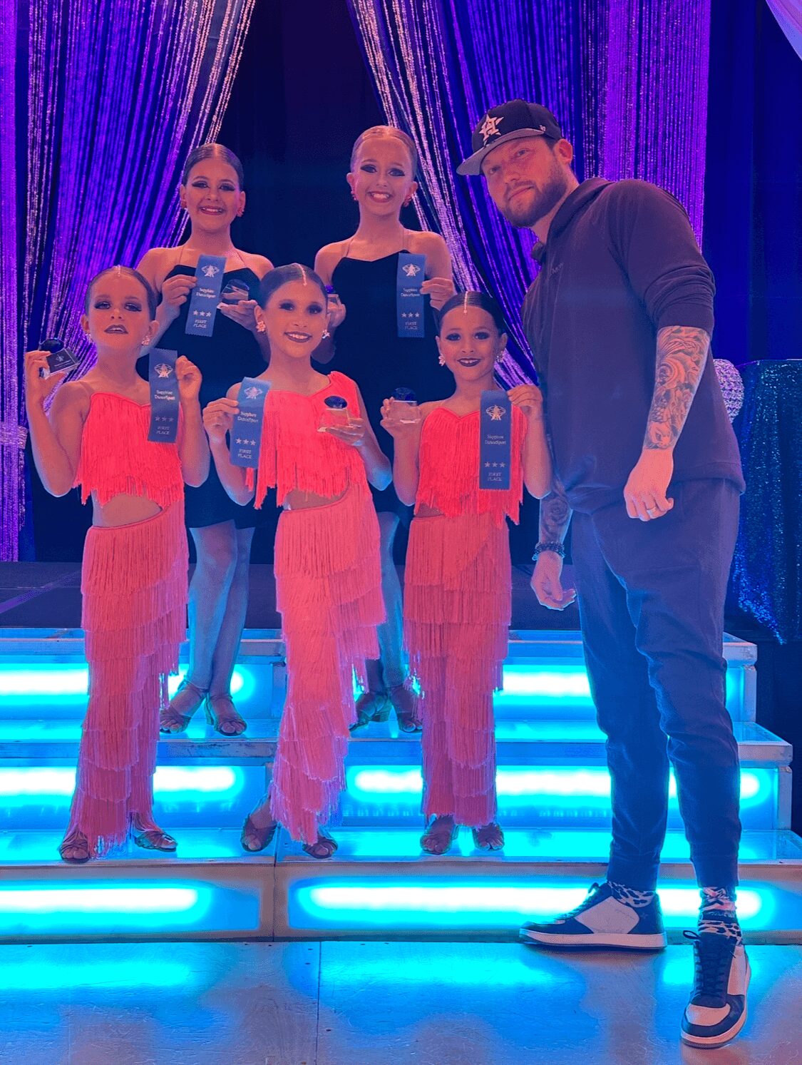 Group of five young girls in pink costumes holding awards, with a man in casual clothing on a brightly lit stage with blue curtains.