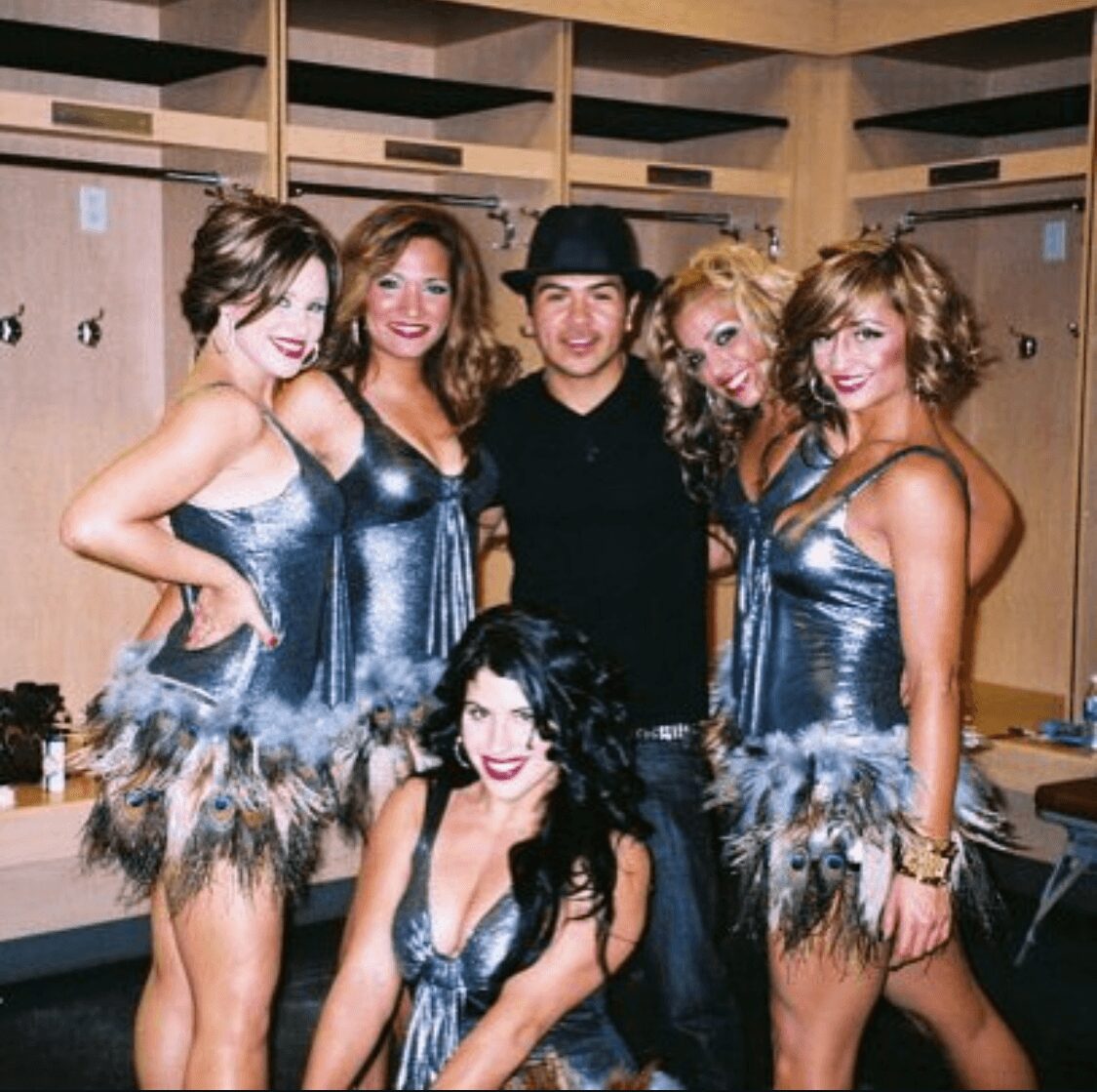 Group of six people in metallic costumes, five women standing and one woman kneeling, in a dressing room.