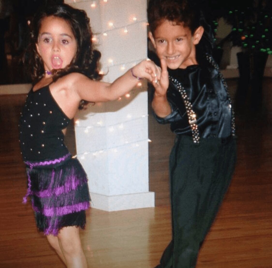 Two children dancing together, holding hands, in a festive setting with string lights and a decorated background.