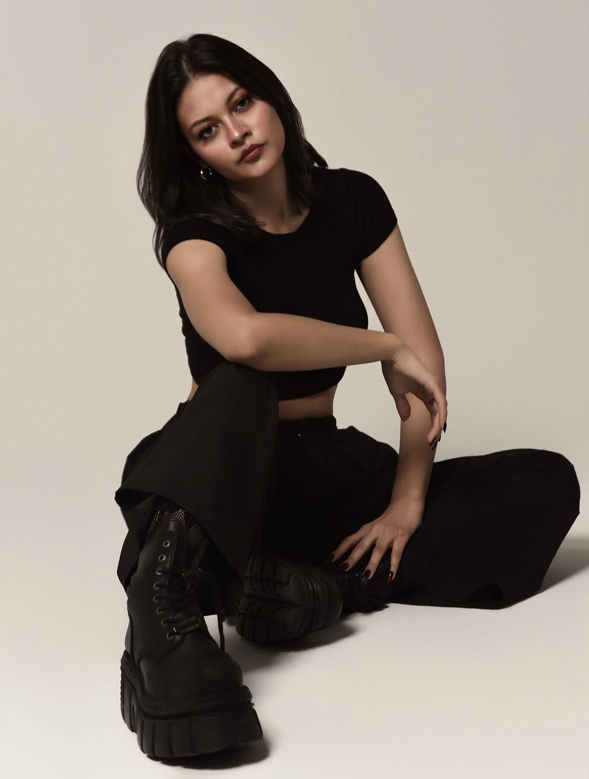 Young woman with dark hair in black clothing sitting on the floor against a plain background.