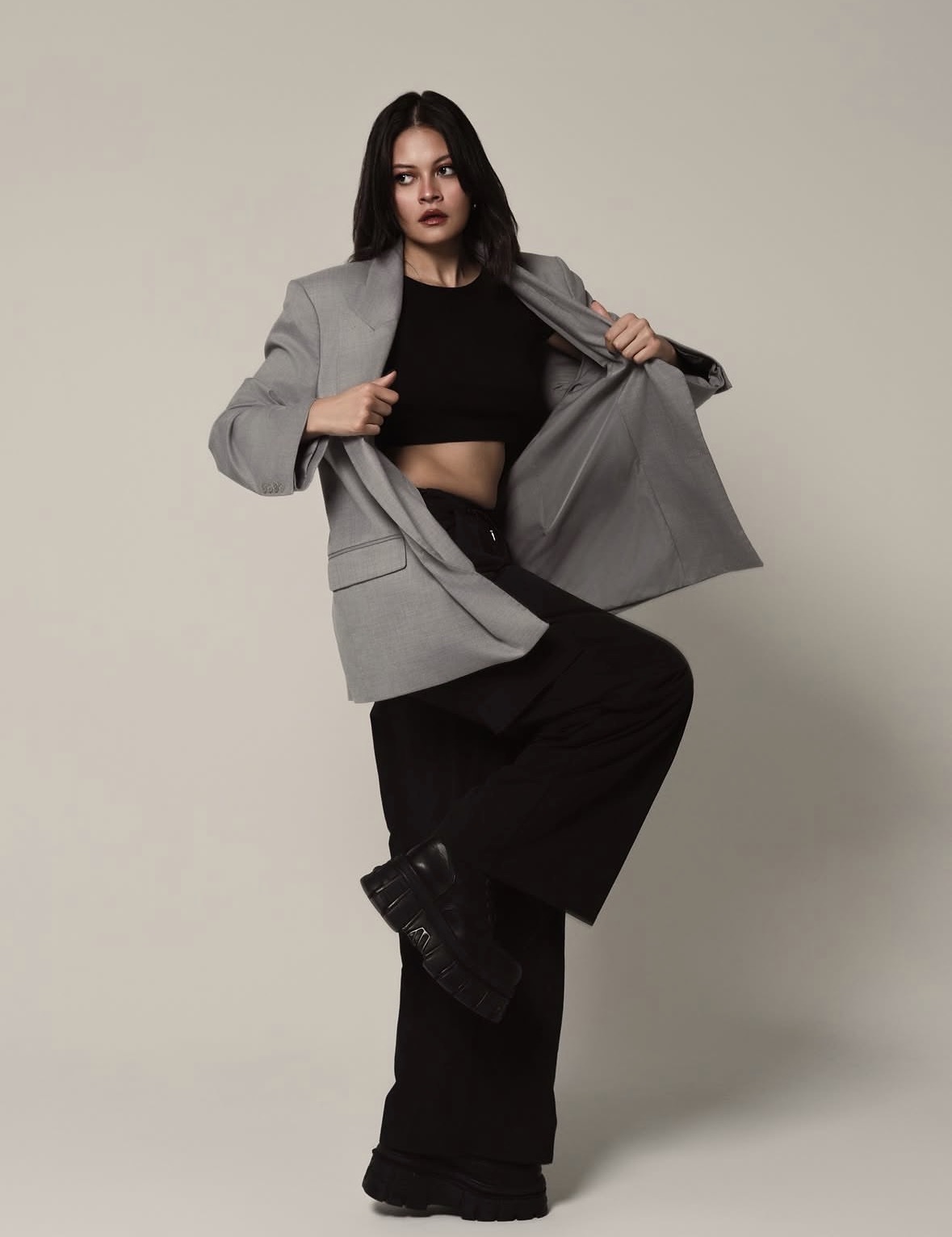Woman in gray blazer and black outfit poses with one leg raised, holding open blazer, against plain background.