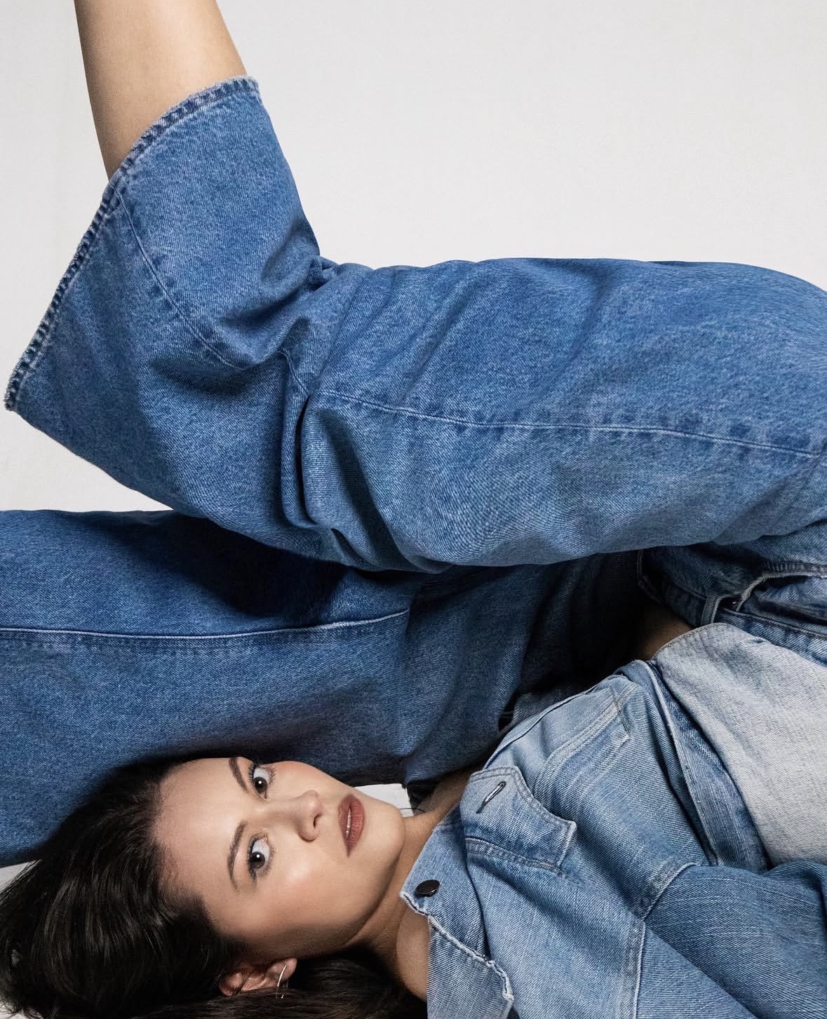 Woman lying down wearing a denim shirt and jeans, looking at the camera, with a plain background.