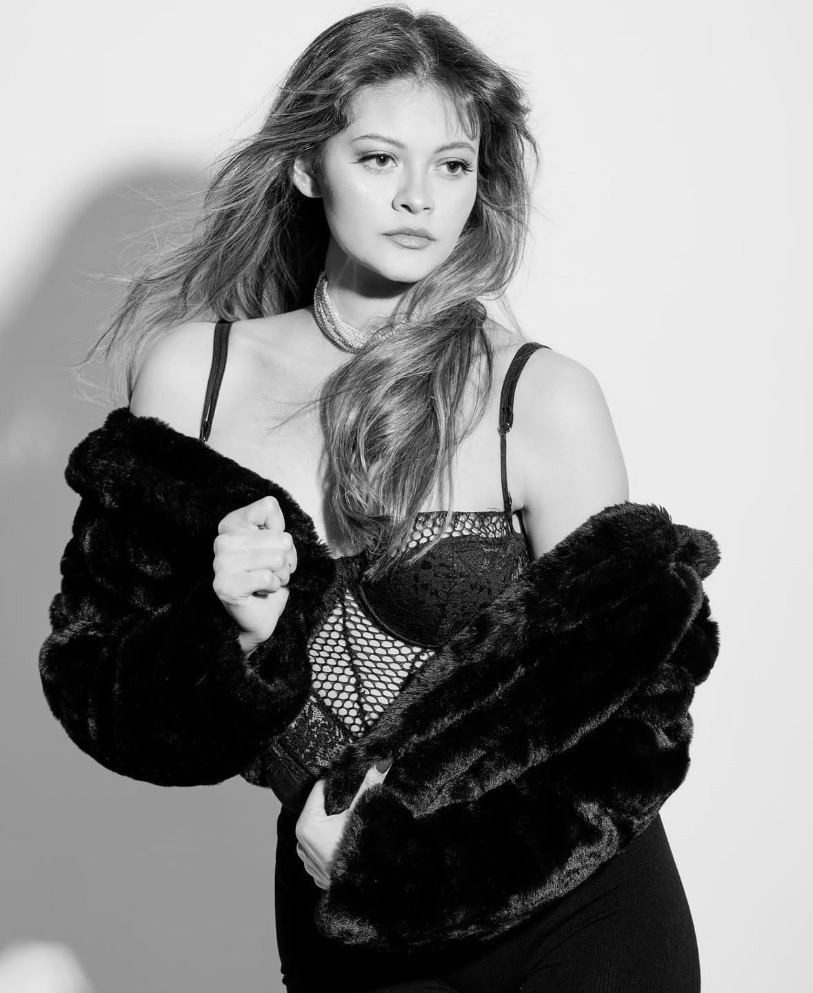 Black and white photo of a woman with long hair, wearing a fur coat and lingerie, looking to the side.