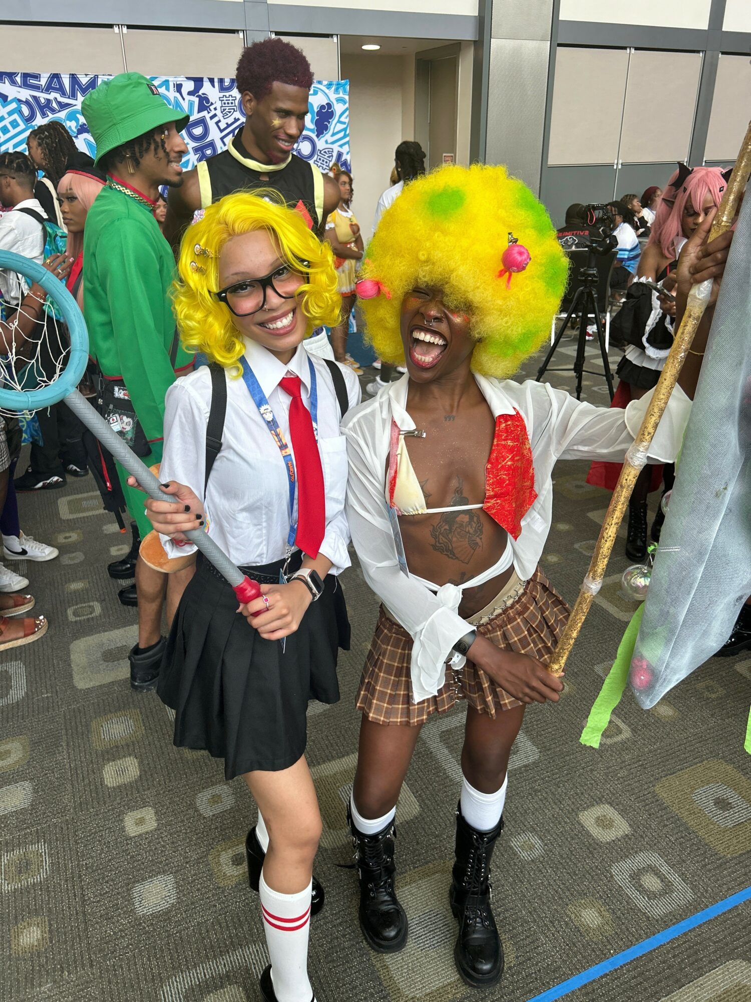 Two people in colorful costumes smiling, one with a yellow wig and the other with glasses and a green hat, at an event.