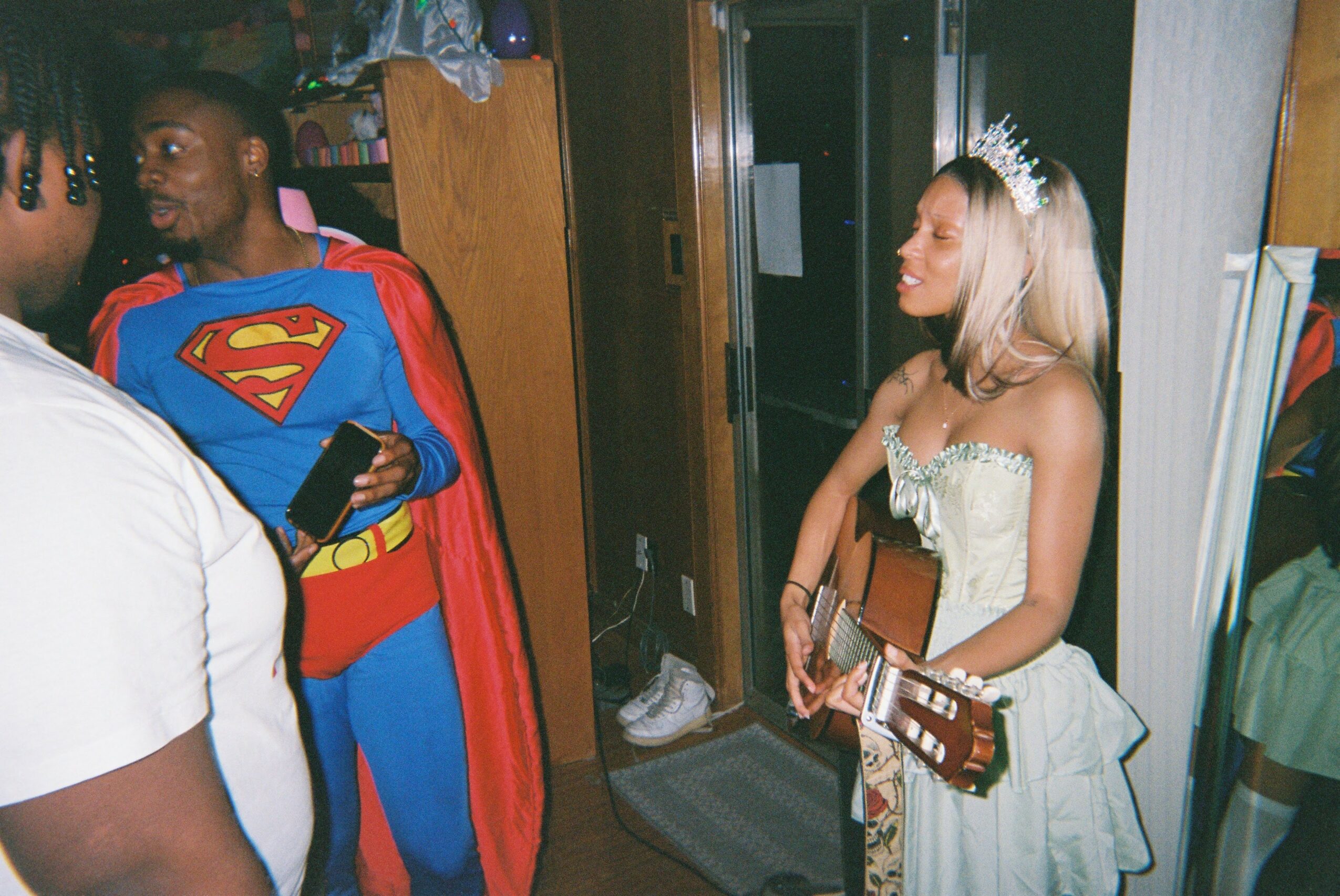 Person dressed as Superman holding a phone, woman in a white dress with a tiara holding a guitar, in a room.