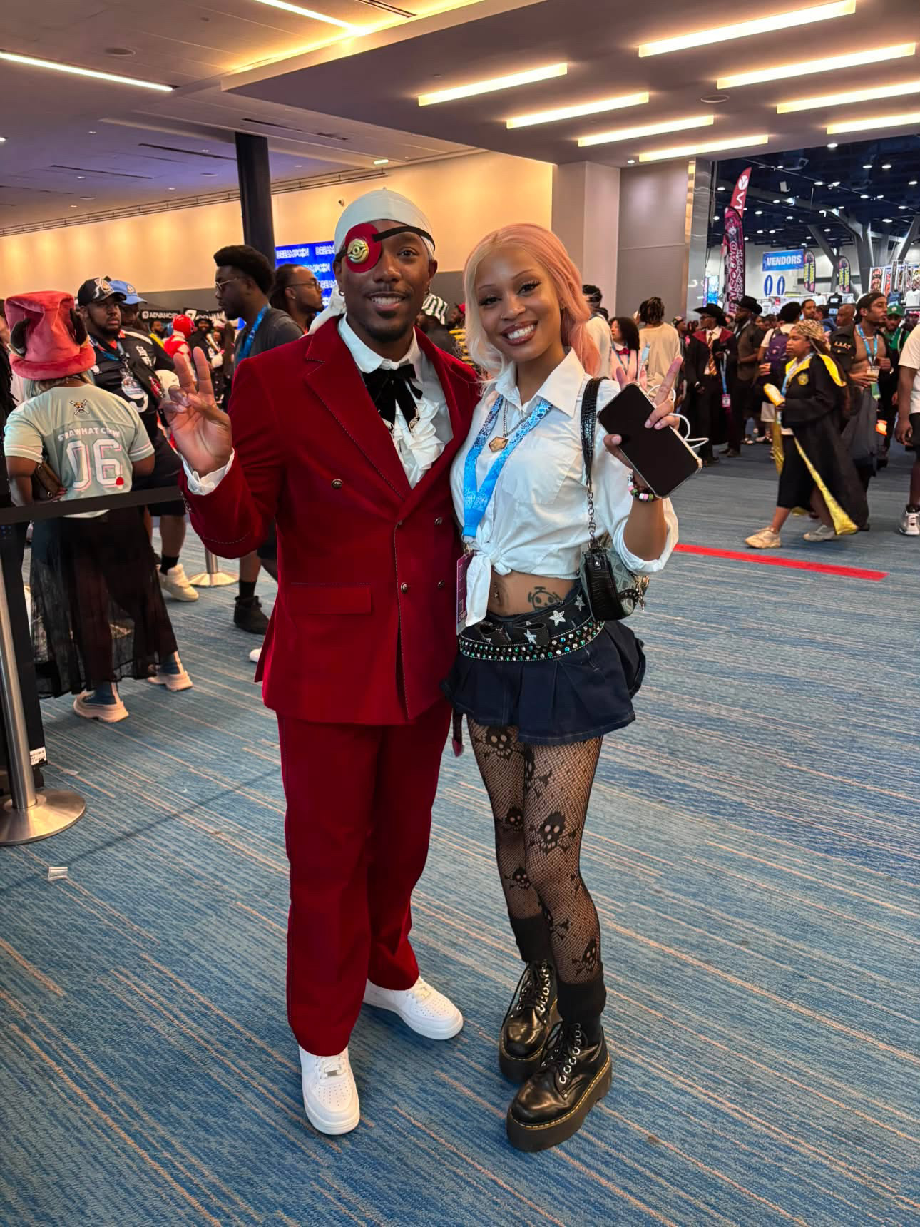 Two people smiling and posing together at an indoor event, with a crowd and bright lights in the background.
