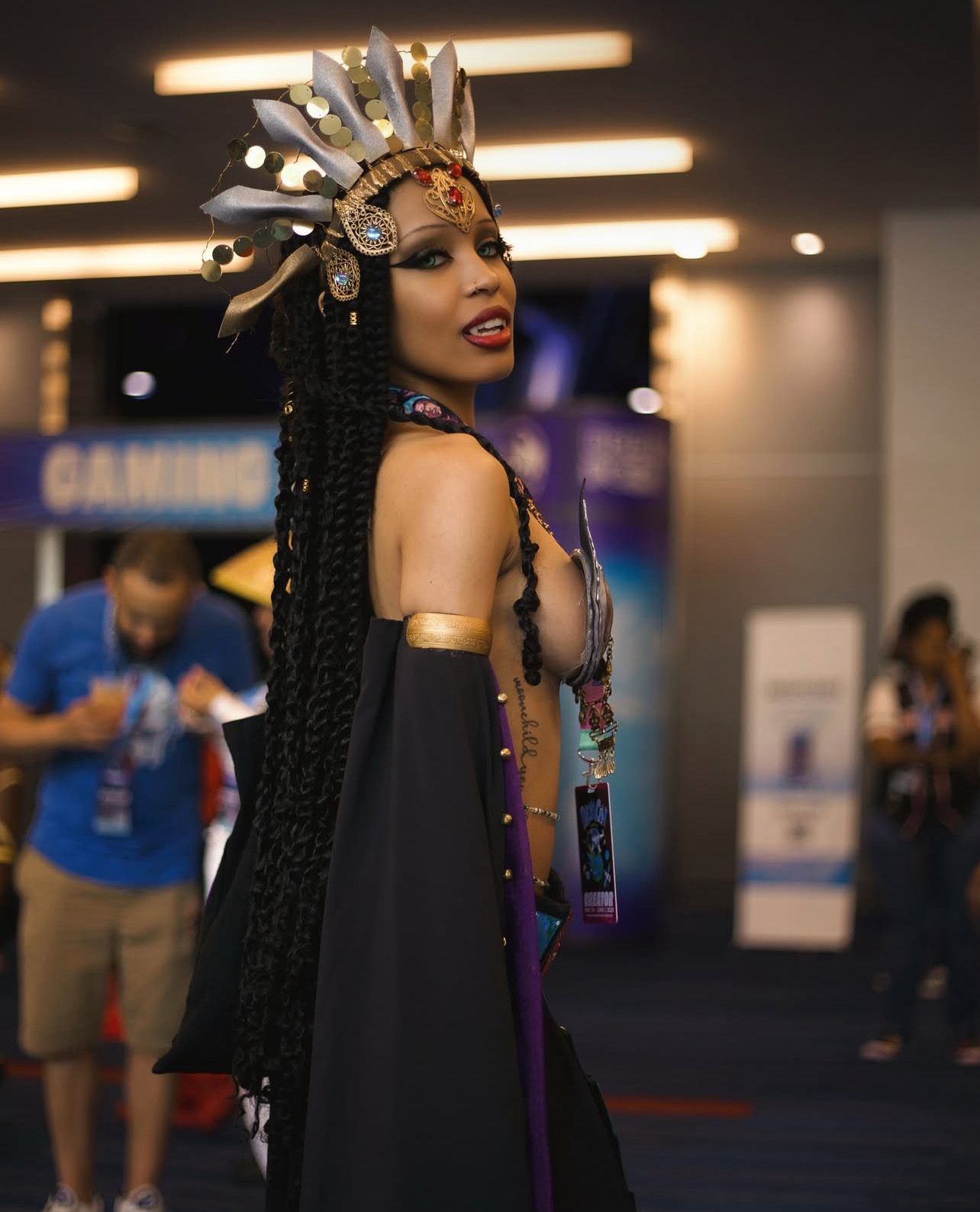 Woman in elaborate costume with headpiece and jewelry at an indoor event, with people and booths in background.