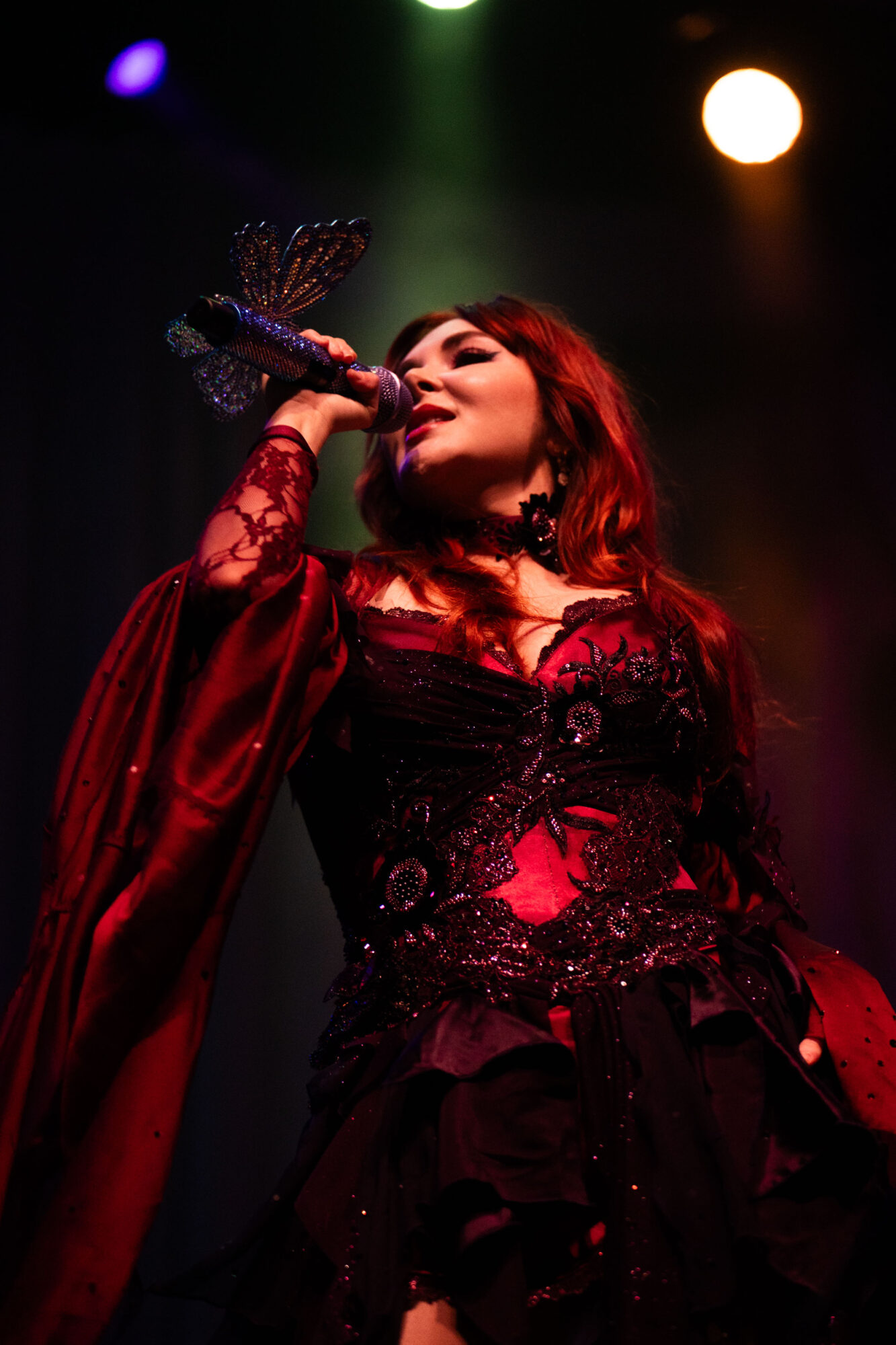 Performer singing into a microphone on stage with colorful lights, wearing a dark, ornate outfit with red hair.