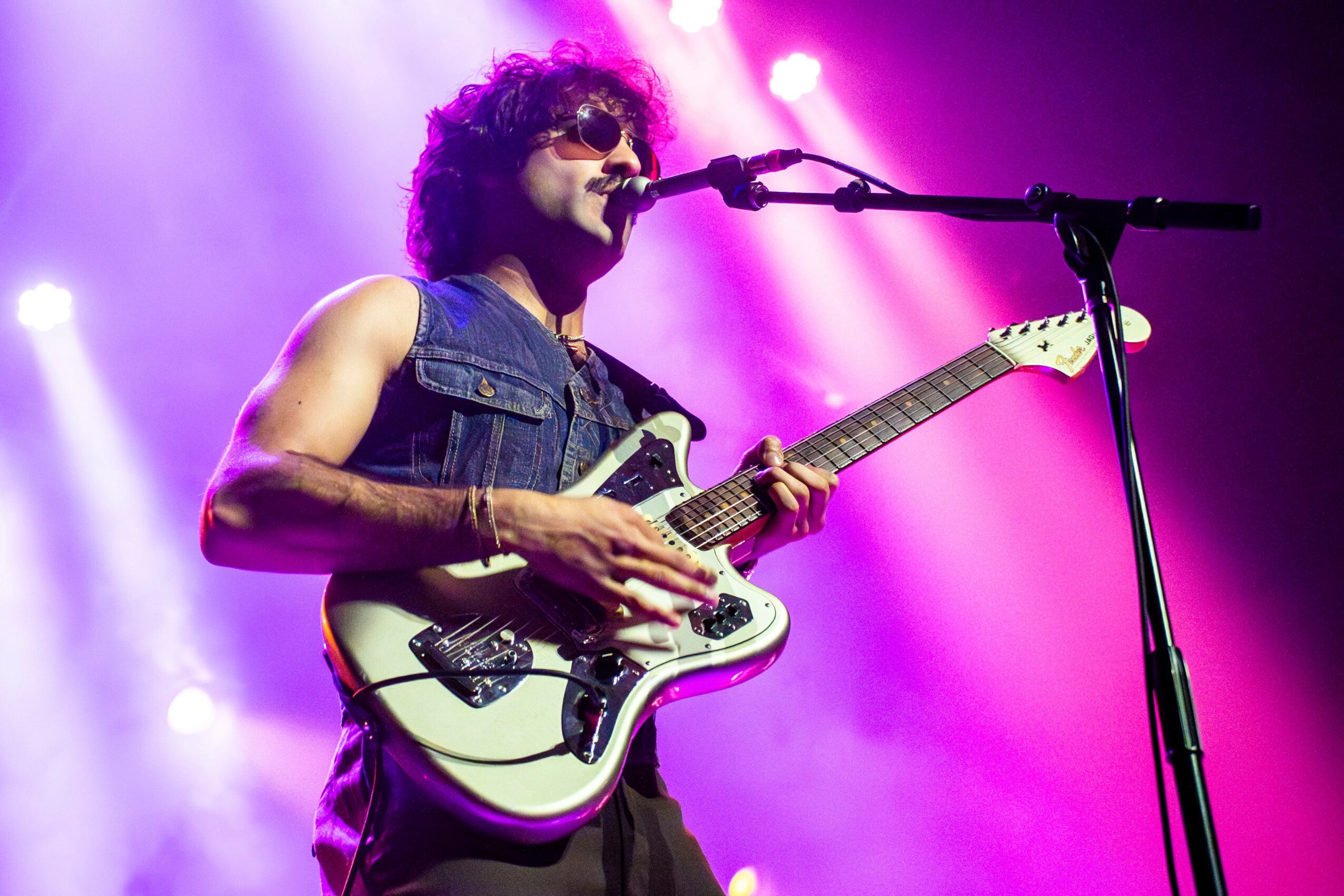 Musician with curly hair and sunglasses playing an electric guitar on stage with purple lighting