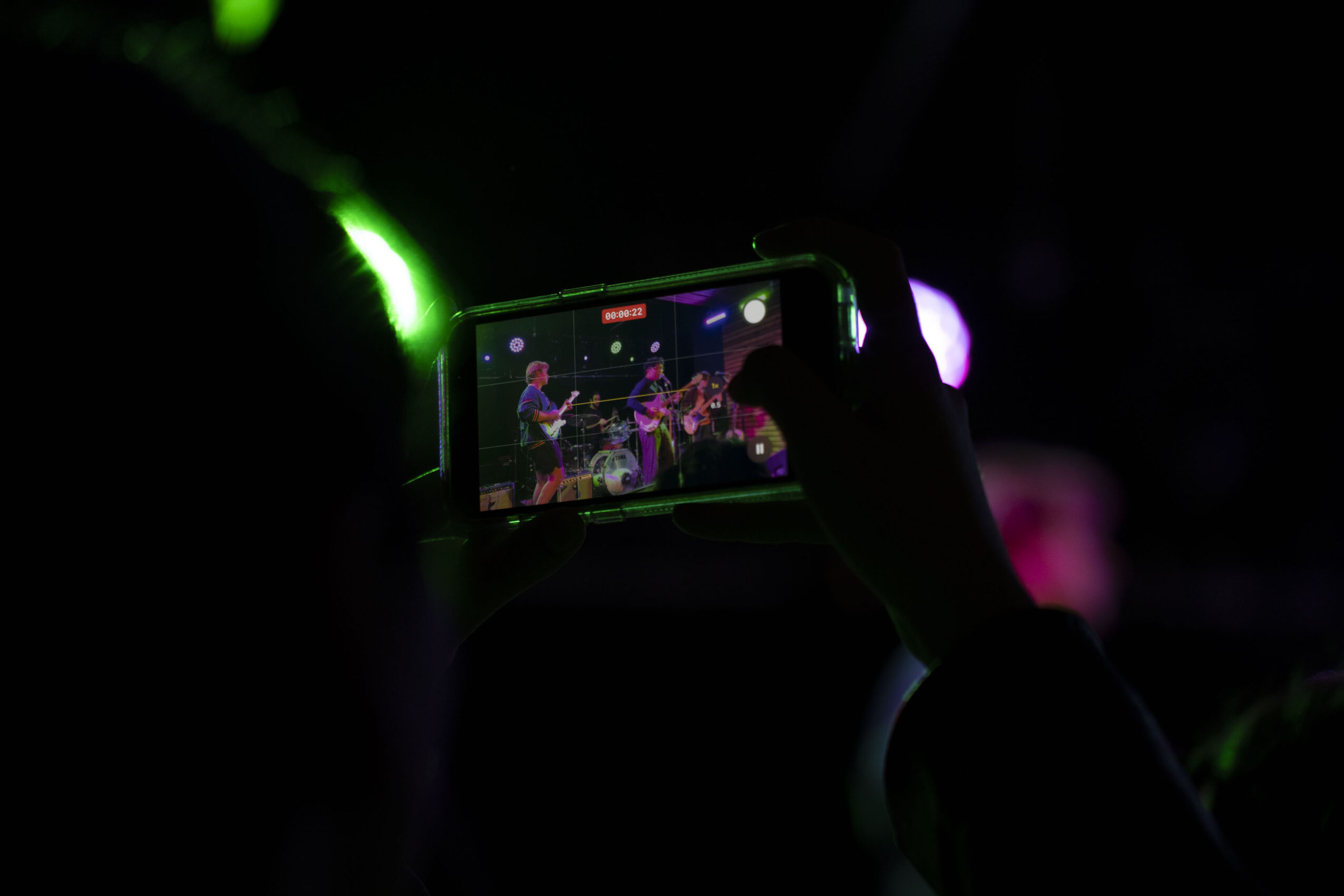 Person recording a live performance on a smartphone in a dark setting with colorful lights, hand visible holding the phone.