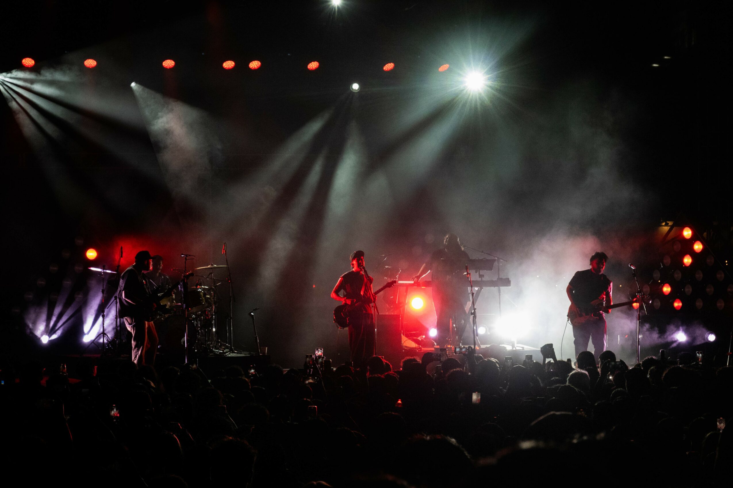 Band performing on stage with bright lights and fog, audience in foreground, colorful stage lighting effects.