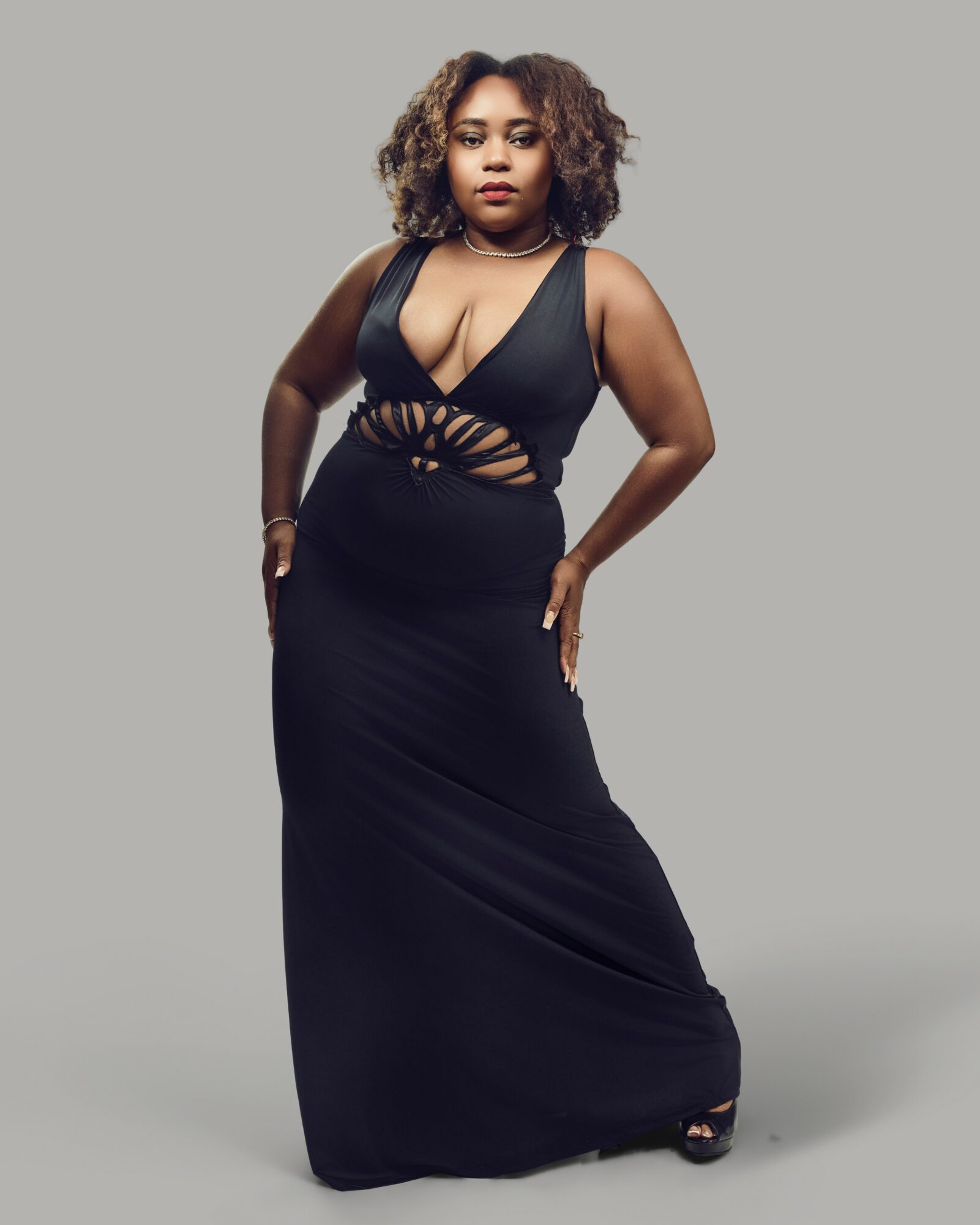 Woman in a black dress with cutouts, standing with hands on hips, facing forward, against a plain gray background.