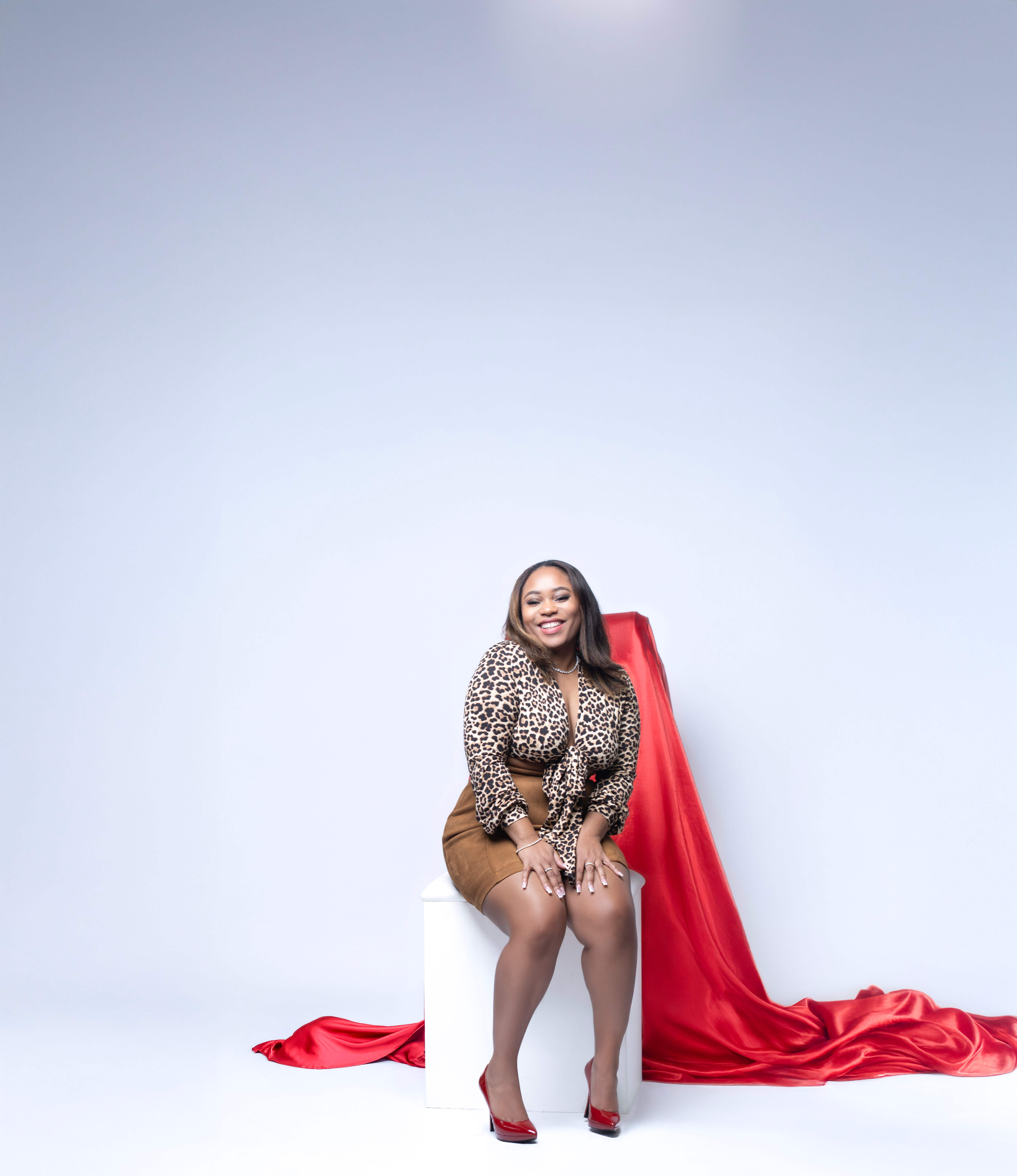 Woman sitting on a white block with a red fabric draped behind her, smiling, against a plain light background.