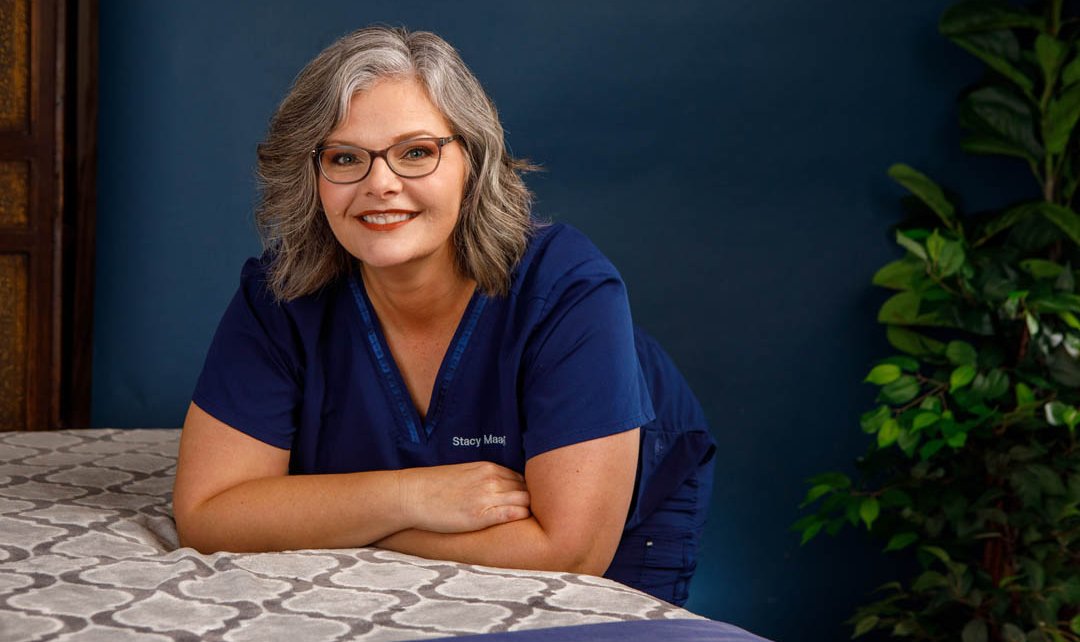 Smiling woman with gray hair and glasses, wearing a blue shirt, sitting at a table with a patterned tablecloth, background with green plant.