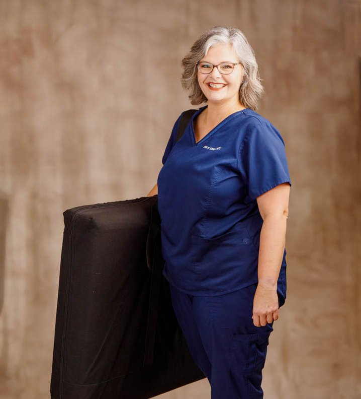 Smiling woman in blue scrubs holding a black cushion, standing against a neutral background.