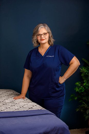 Woman in navy scrubs with glasses standing beside a bed, hand on hip, dark blue background, plant nearby.