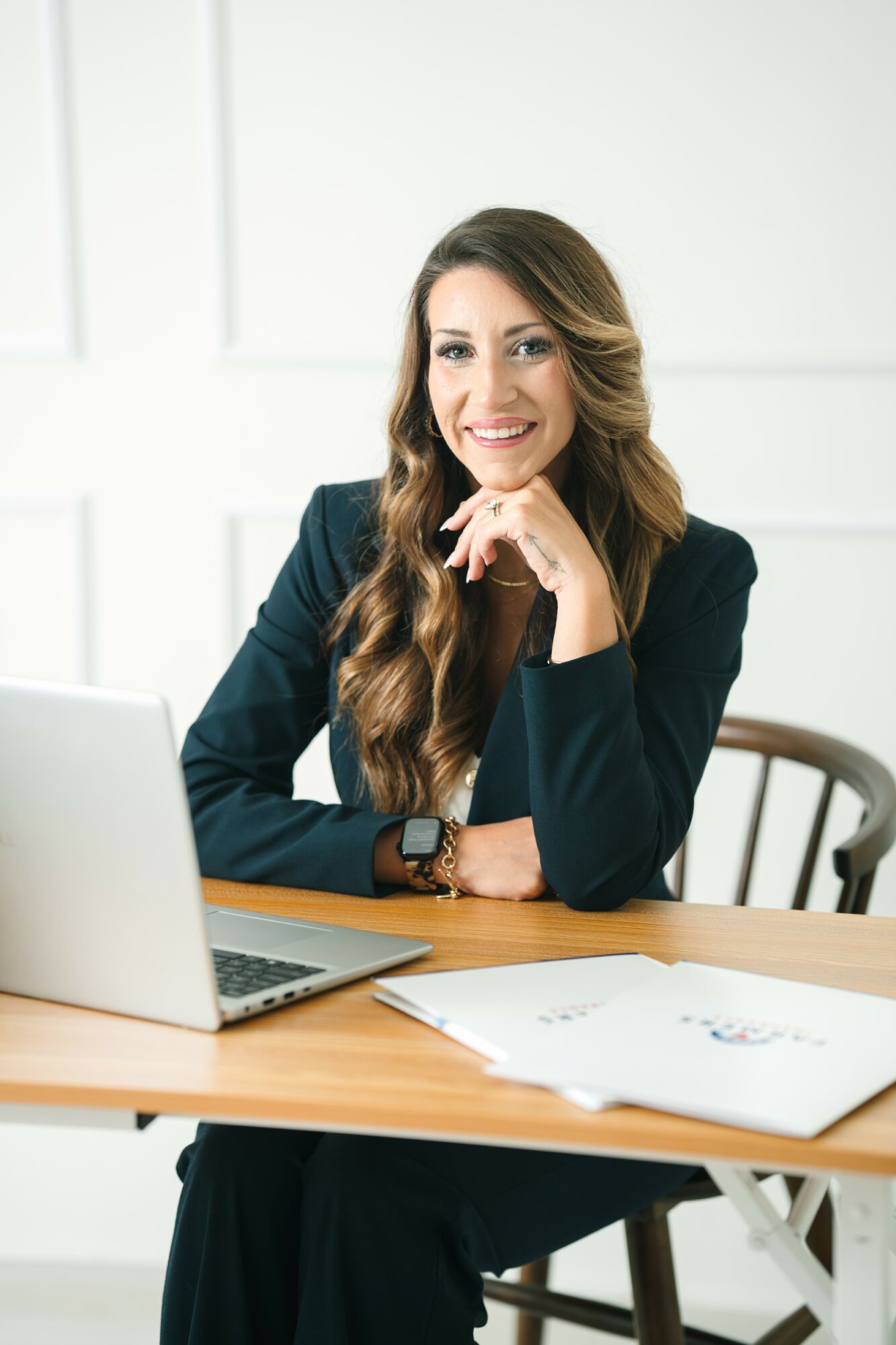 Smiling woman with long wavy hair sitting at a desk with a laptop and papers, in a bright office setting.