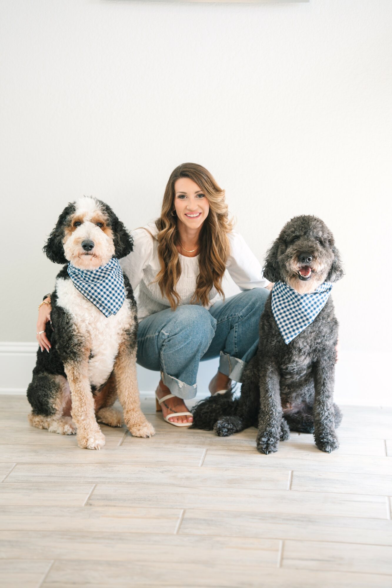 Woman crouching between two dogs with bandanas, smiling, against a plain white background.