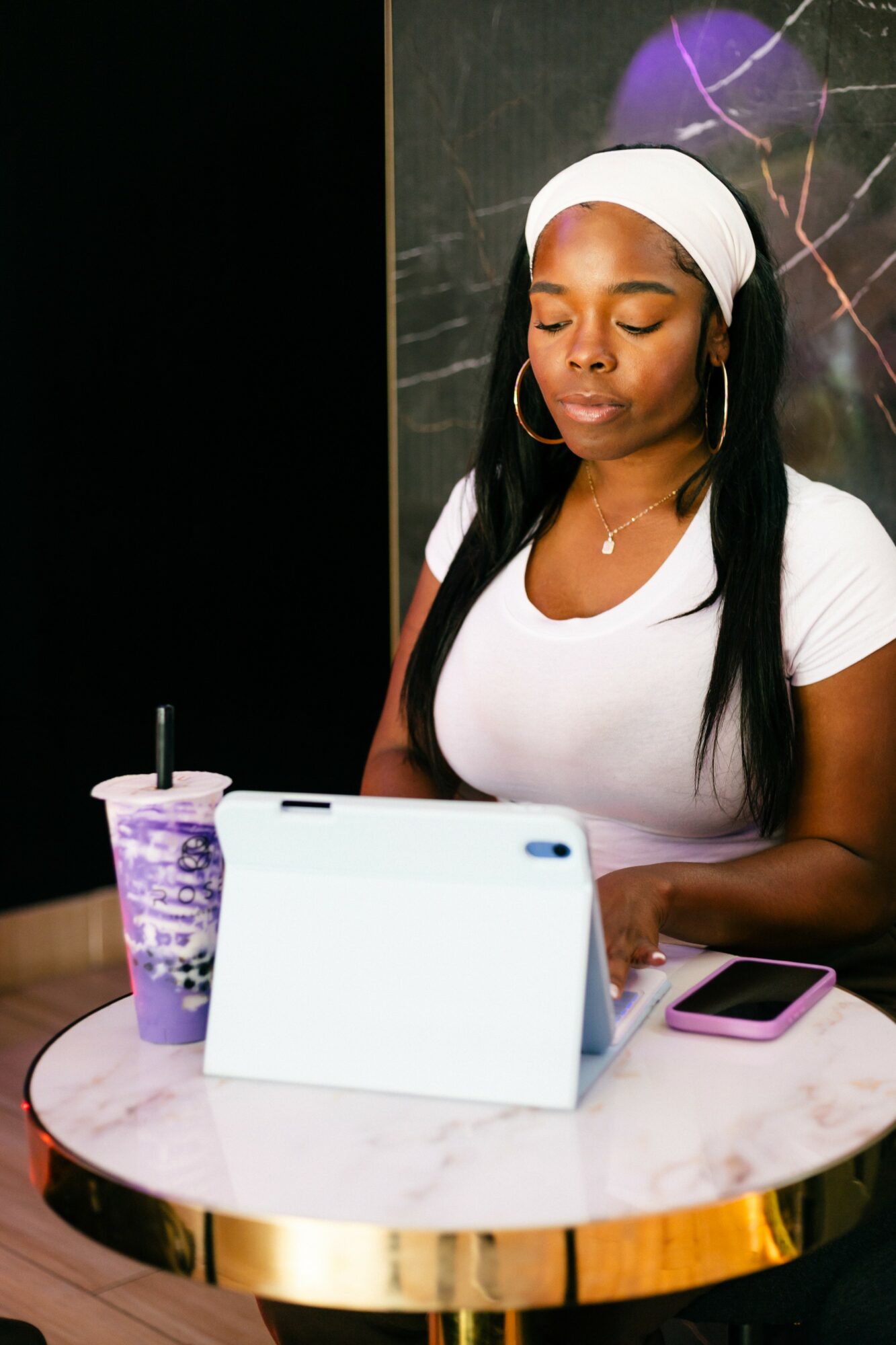 Woman with white headband and shirt sitting at a table with a tablet, phone, and drink, eyes closed.
