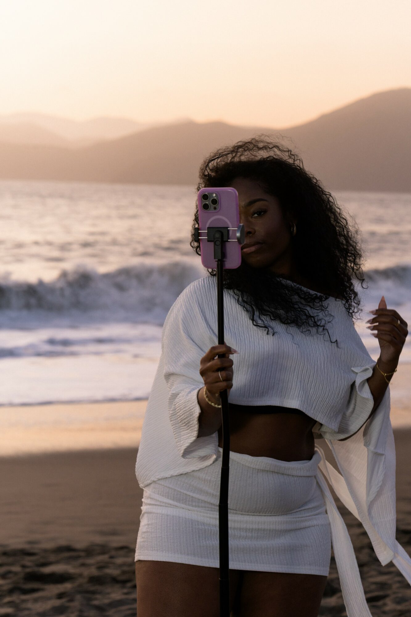 Woman with curly hair taking a selfie on the beach at sunset, ocean waves in background, wearing white outfit.