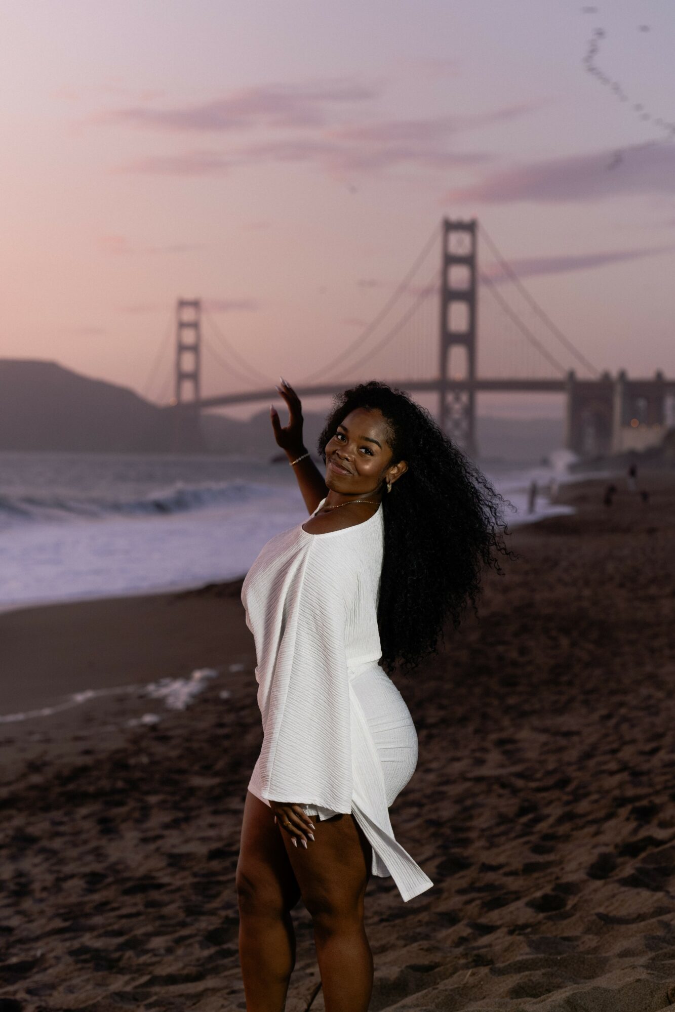 Woman with long curly hair in white dress on beach at sunset, bridge in background