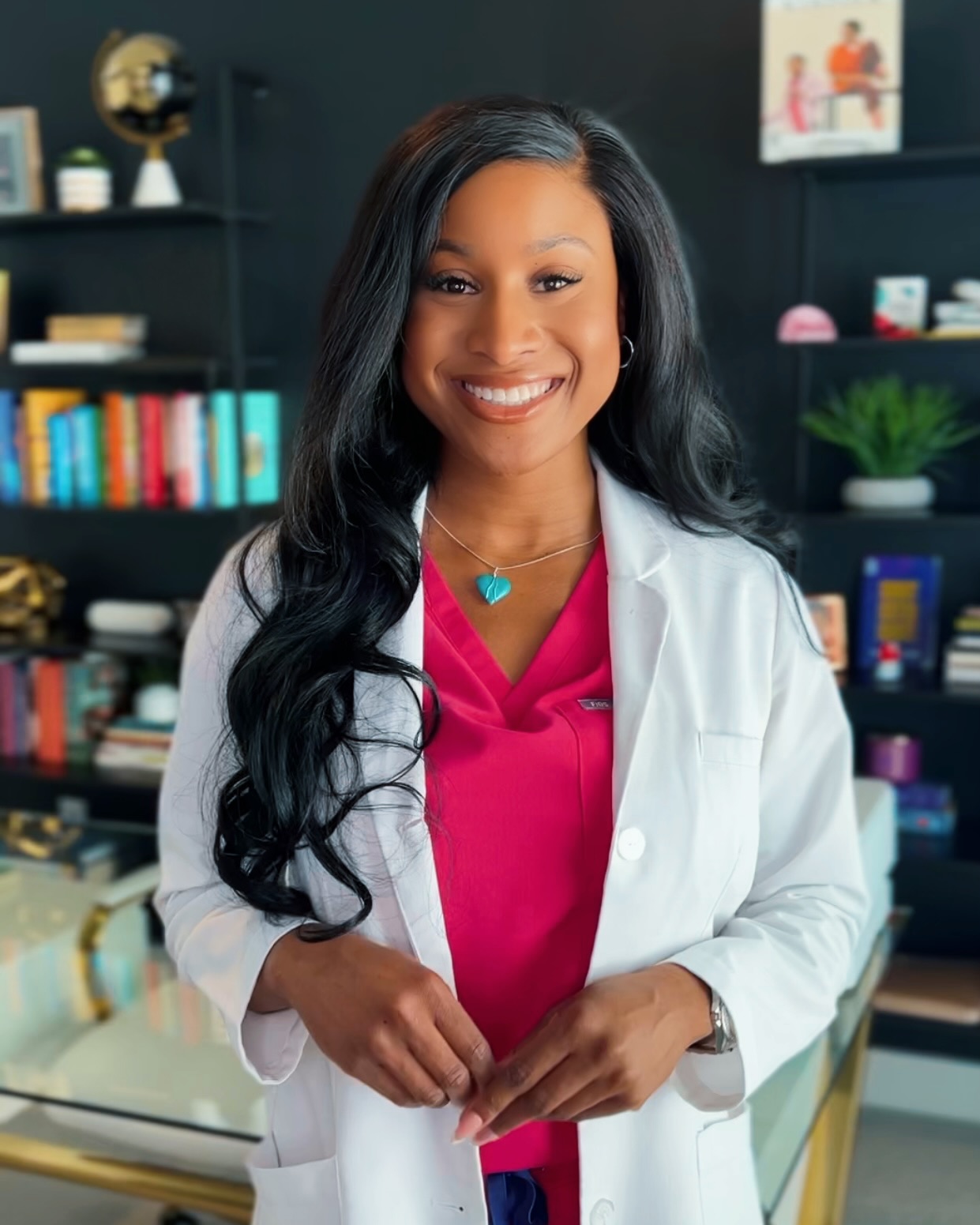 Smiling woman in a white lab coat and pink scrubs standing in a modern office or clinic with books and plants behind her.