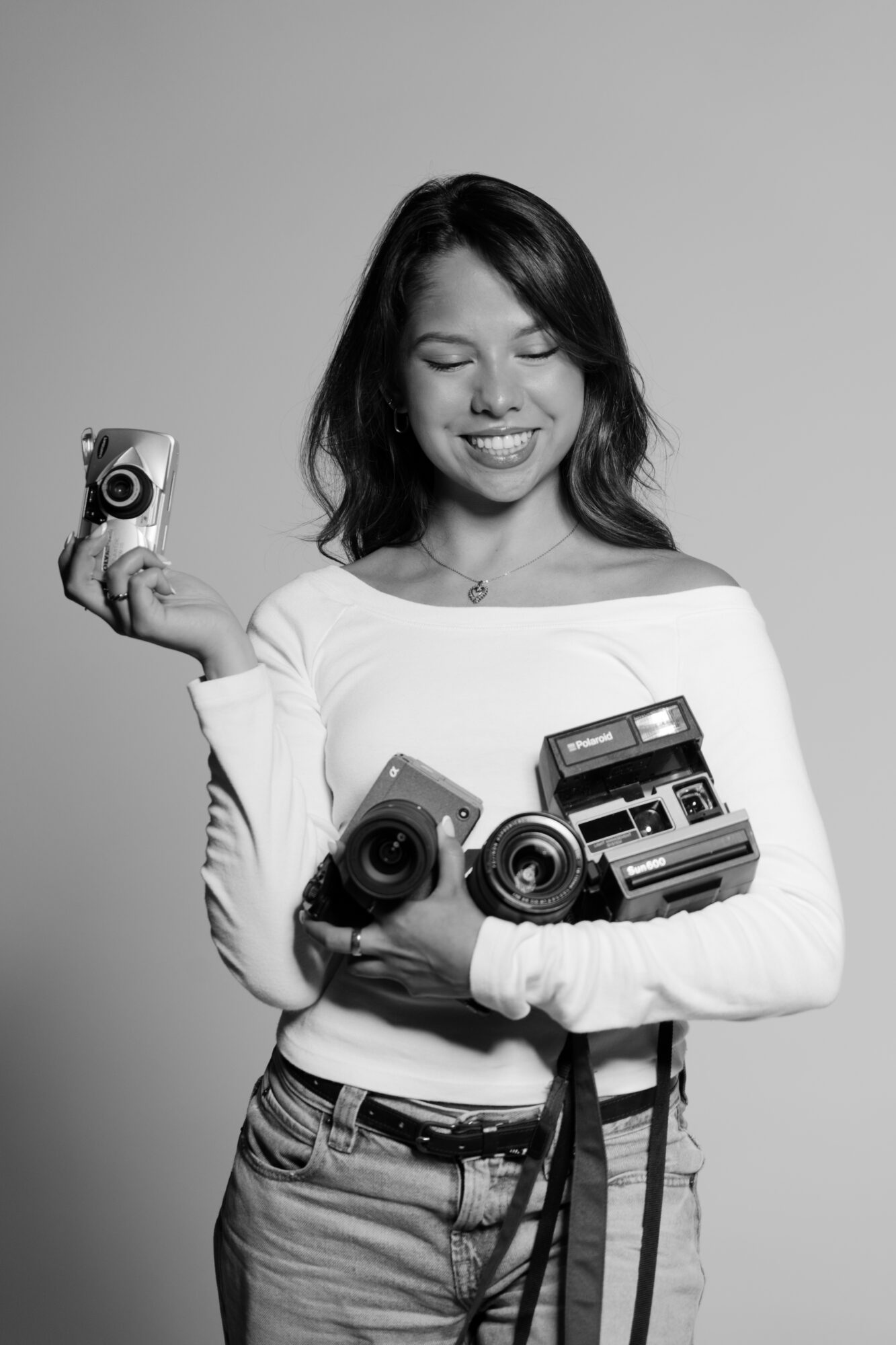 Young woman smiling, holding a camera in her right hand and several cameras in her left arm, wearing a white top and jeans.