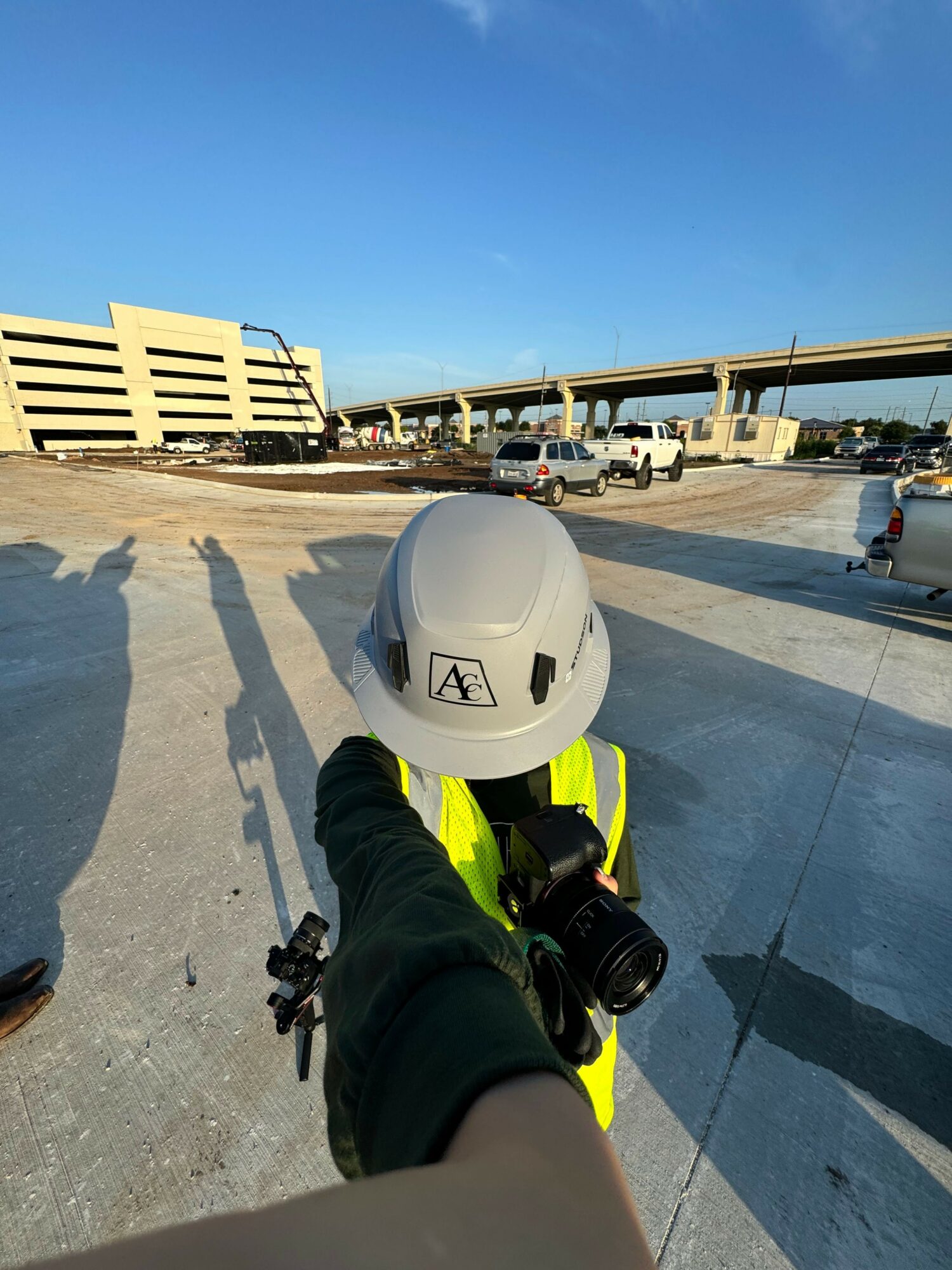 Person in safety vest holding a white helmet and camera on a sidewalk near a body of water with buildings in the background.
