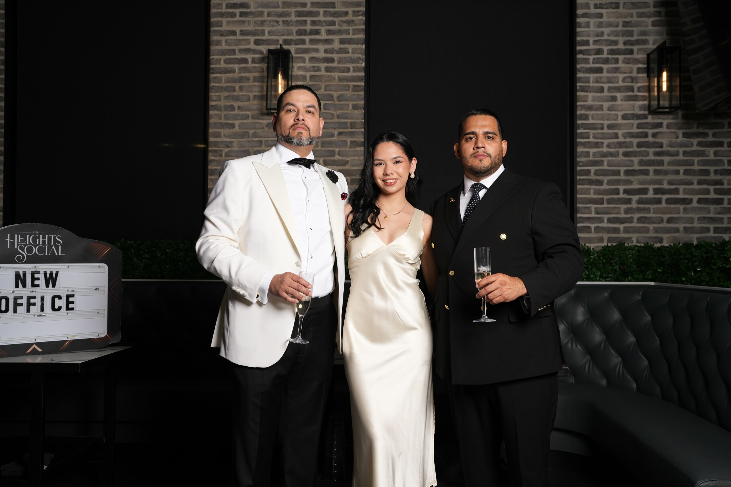 Three people in formal attire holding drinks, standing in front of a brick wall at night.