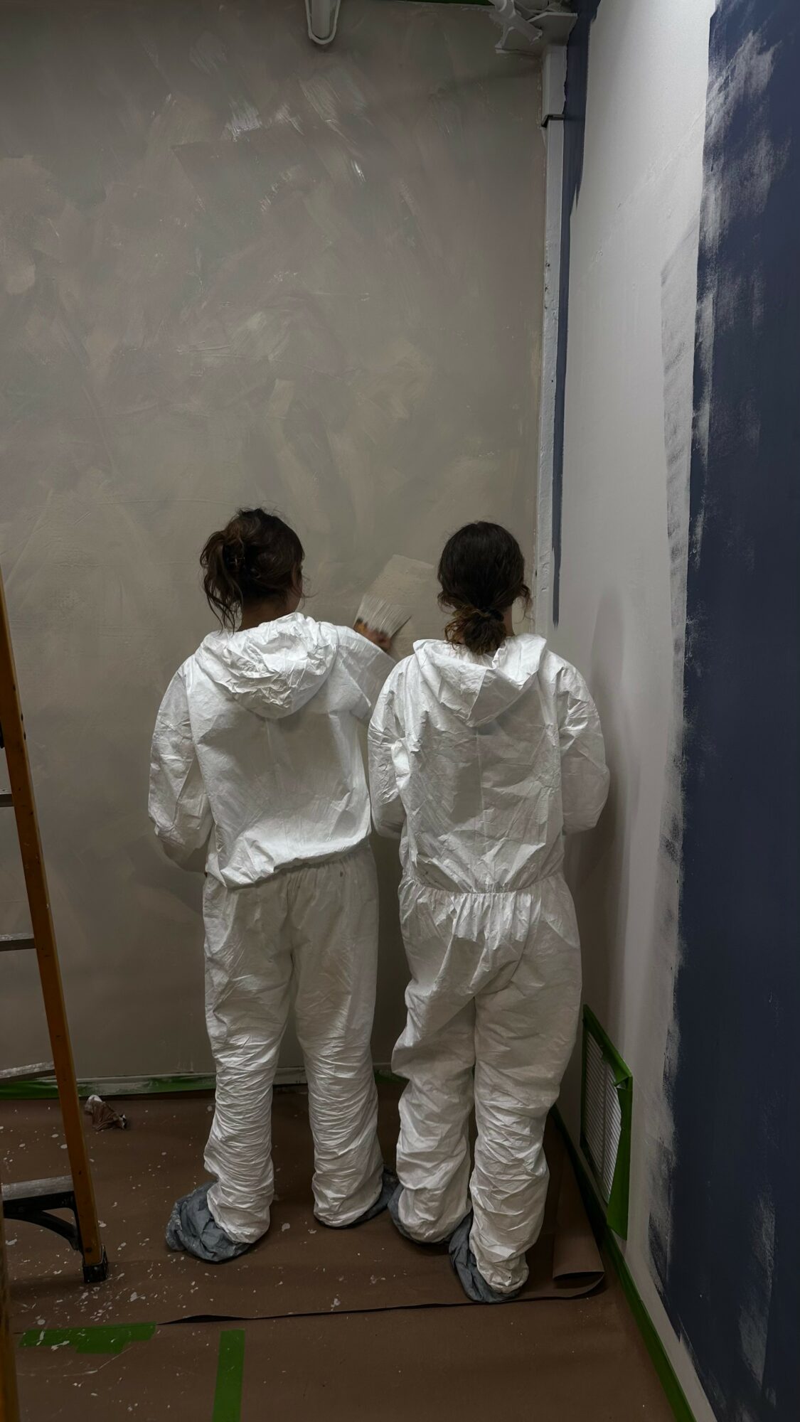 Two people in white protective suits stand with their backs to the camera, facing a wall with a painted sky scene.