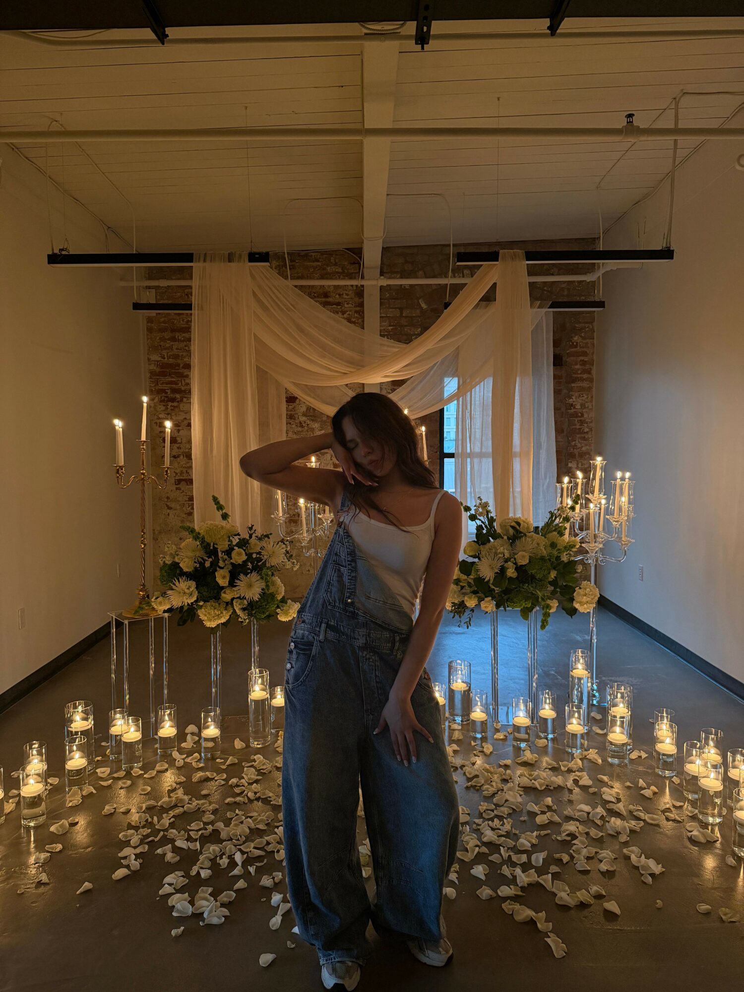 Woman in a white top and blue jeans standing in a decorated indoor space with flowers and candles.