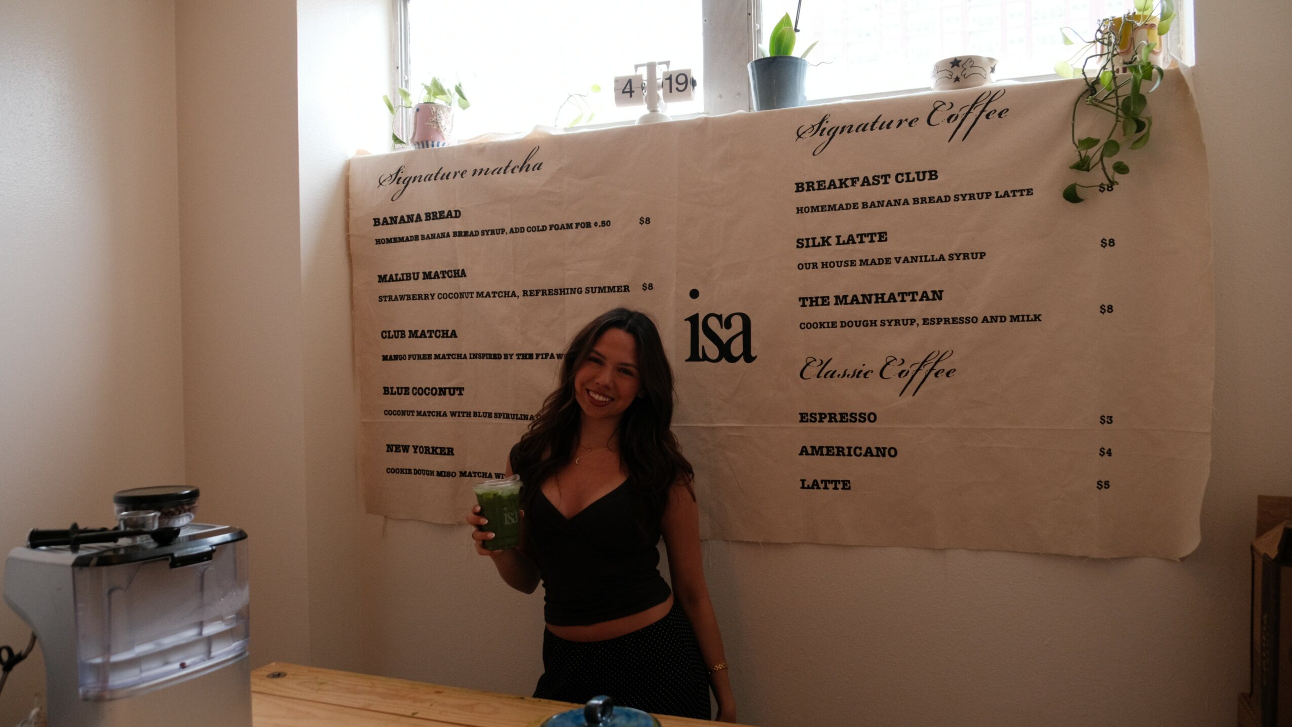 Woman holding a drink standing in front of a large menu board on a wall, with sunlight coming through windows above.