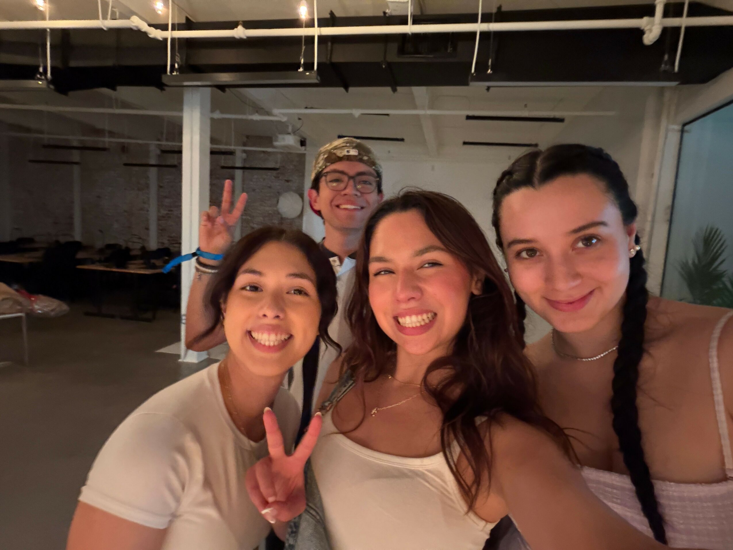 Three women smiling and taking a selfie indoors with a person in the background, some people and objects visible.