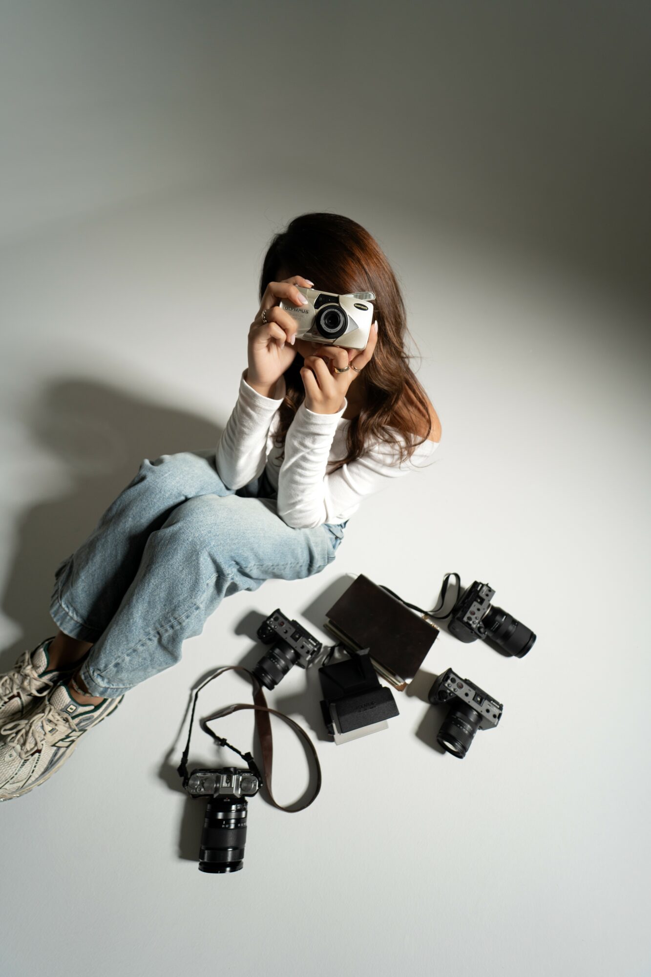 Person sitting on the floor holding a camera, surrounded by multiple cameras and camera accessories, against a plain background.