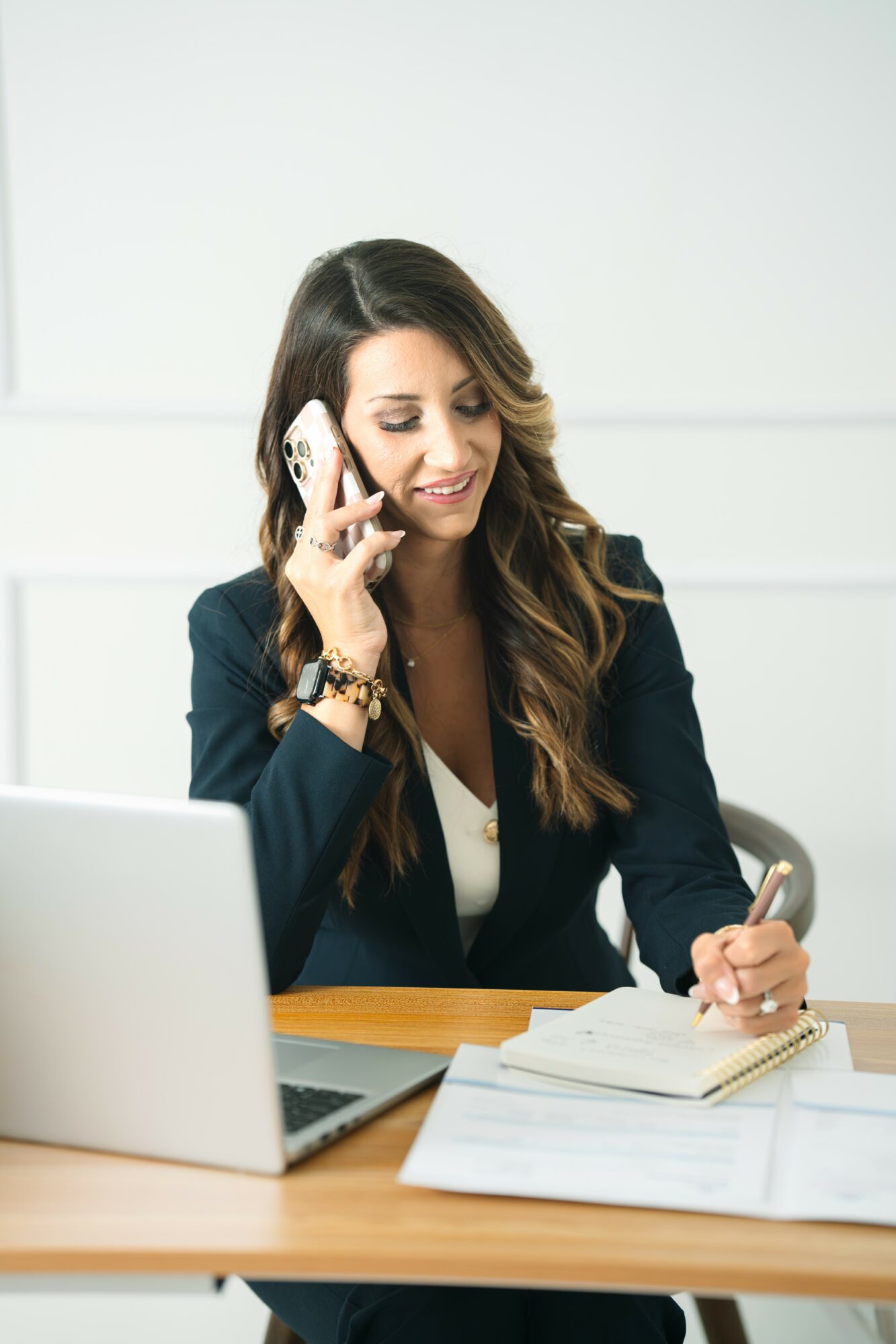 Woman with long hair, wearing a dark blazer, talking on a phone, sitting at a desk with a laptop and papers.