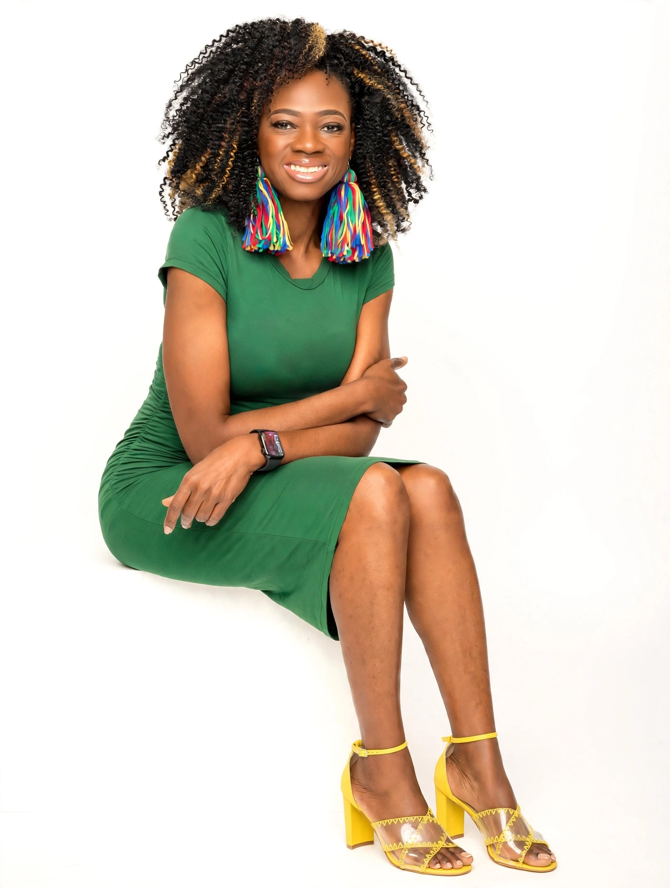 Woman with curly hair wearing a green dress, colorful earrings, yellow heels, sitting with crossed legs, smiling.