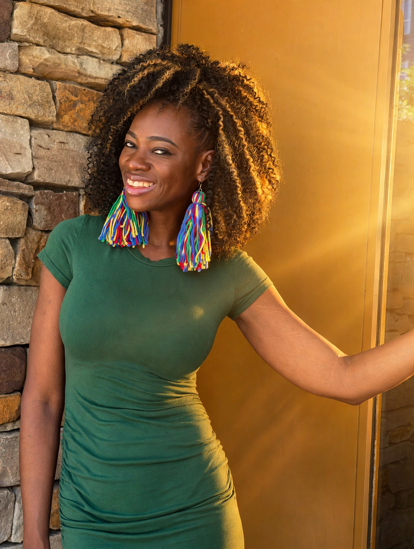 Smiling woman with curly hair wearing colorful earrings and a green dress, standing indoors near a stone wall and golden background.