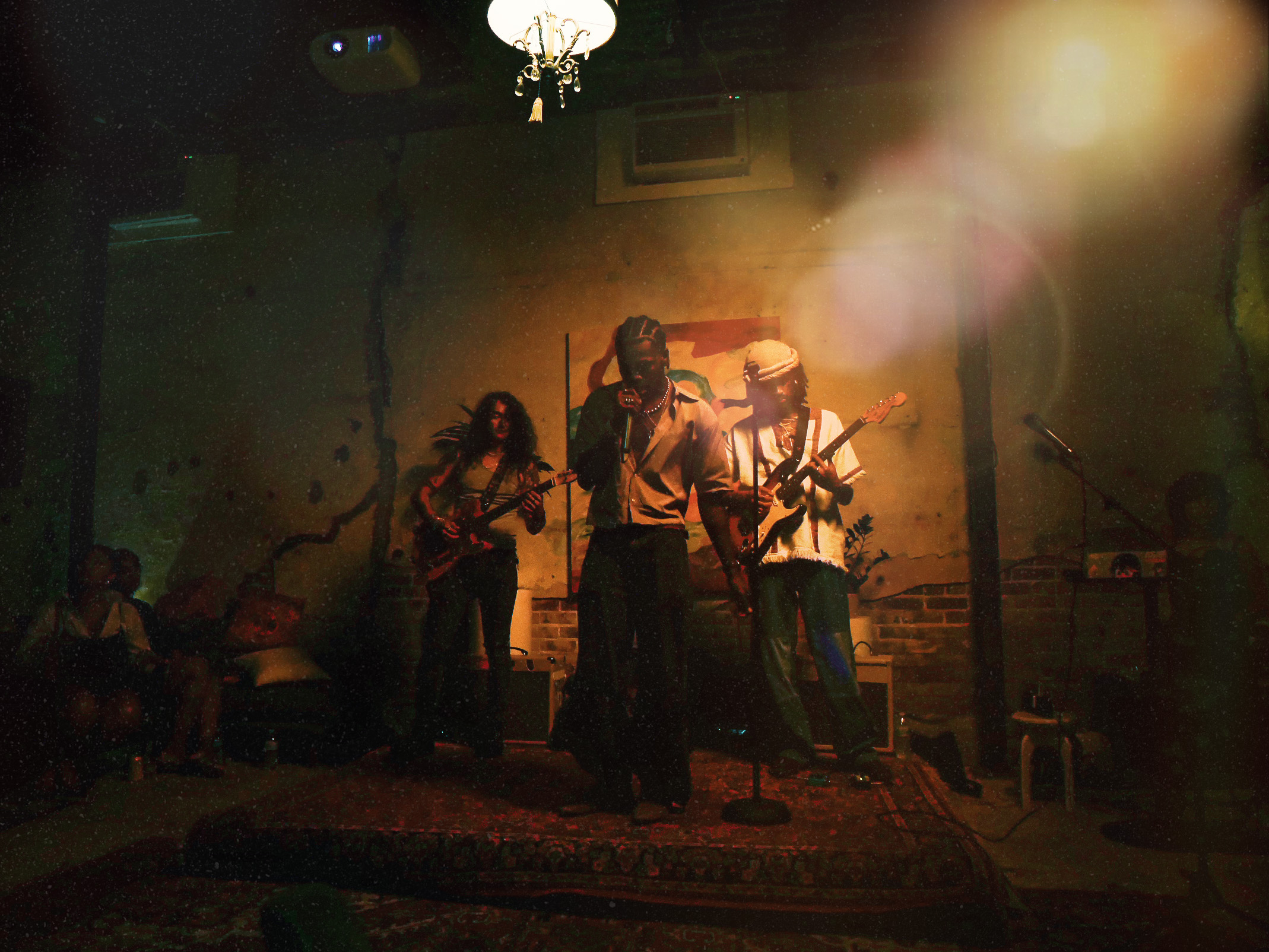 Four musicians perform on a dimly lit stage with colorful lighting and a patterned rug, playing guitars and singing.