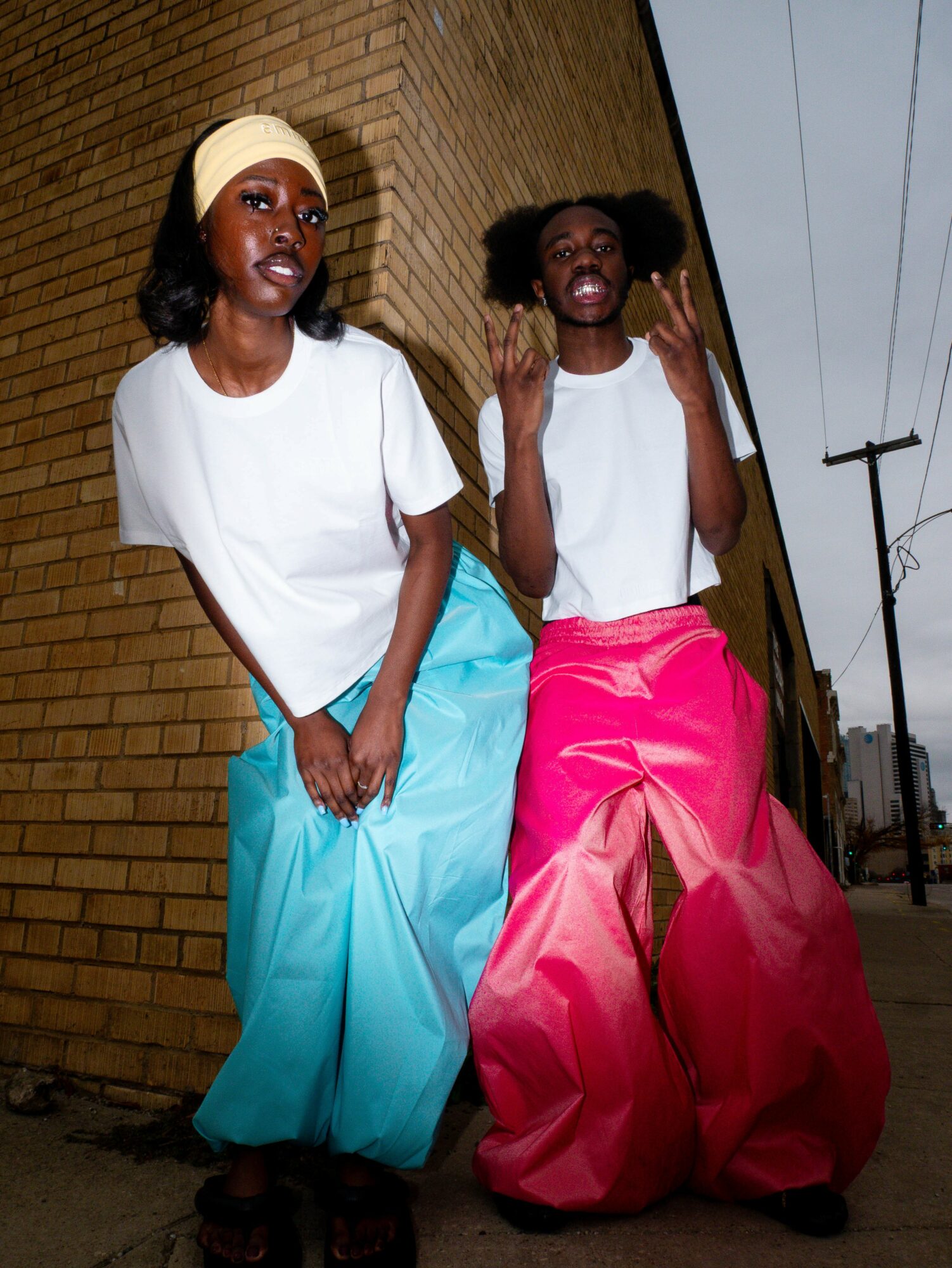 Two women with large pants and white tops standing against a brick wall on a street, one making a peace sign.
