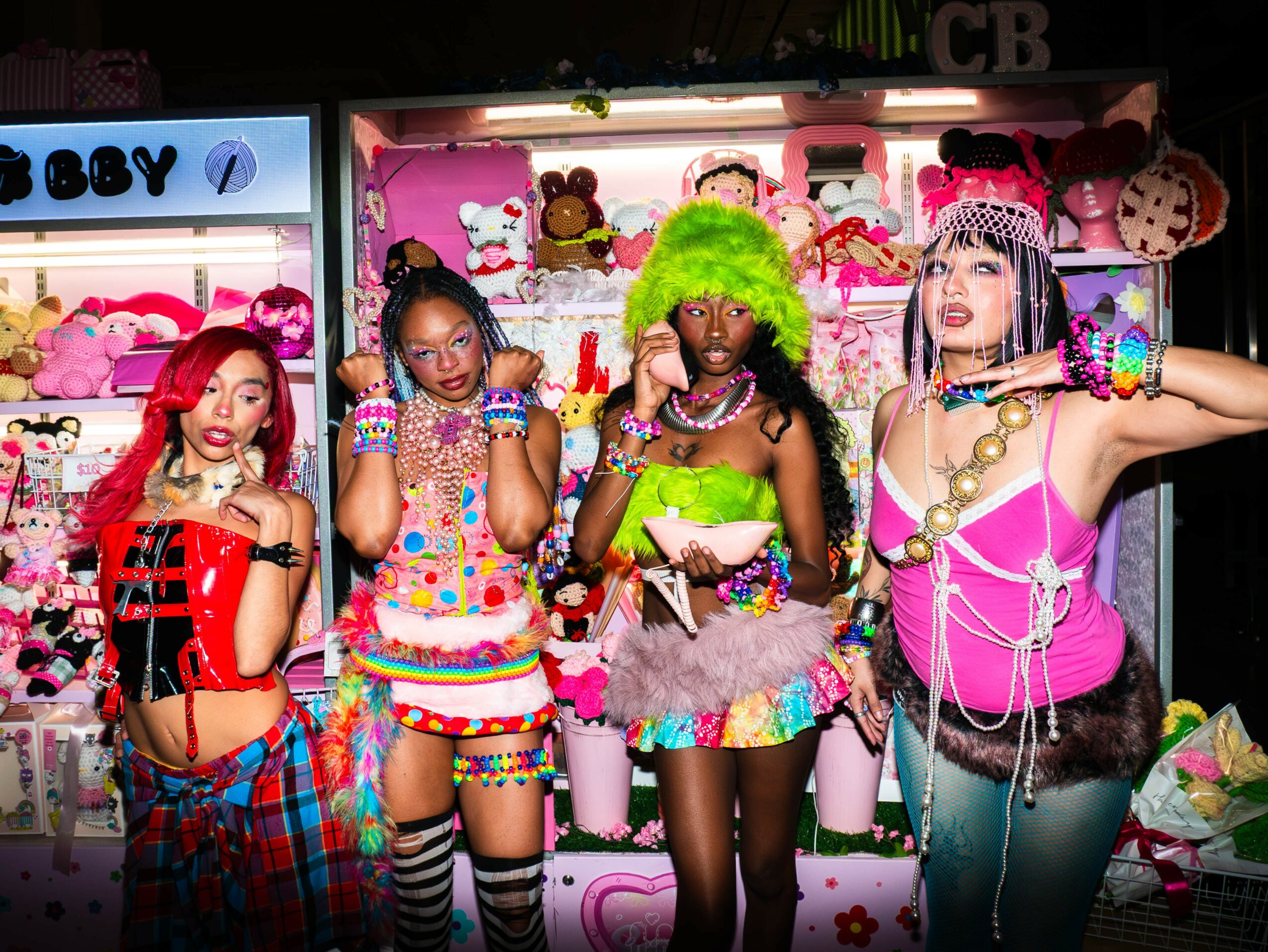 Four women in colorful outfits pose in front of a pink display with plush toys and accessories, smiling and making gestures.