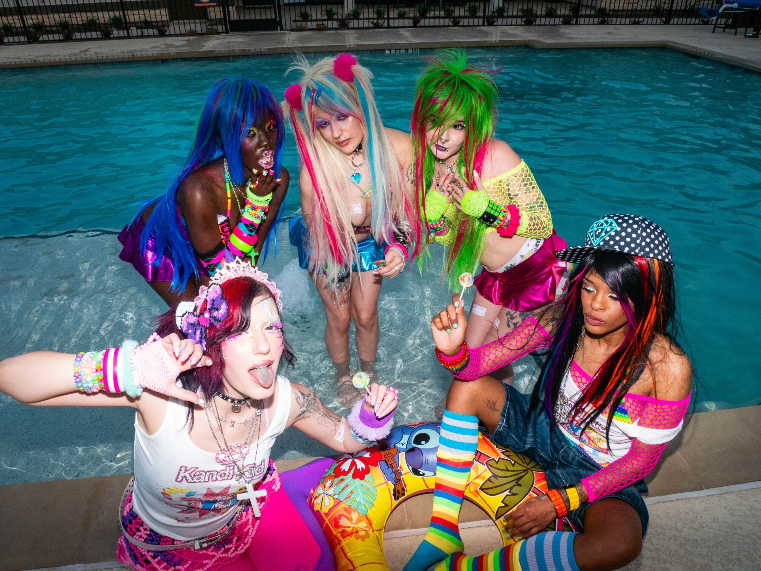 Five people in colorful costumes and wigs by a swimming pool, posing playfully with water in the background.