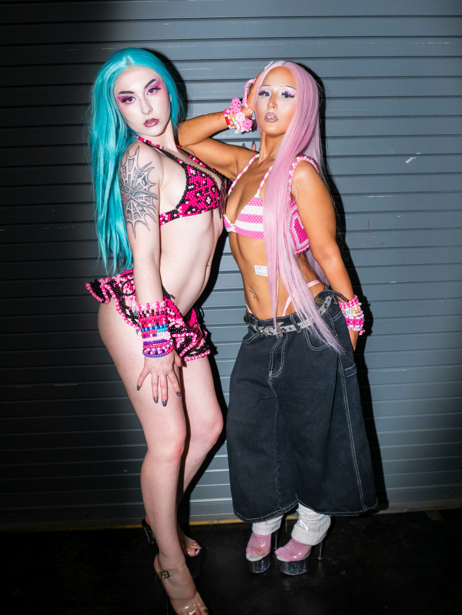 Two women with colorful hair and makeup pose against a gray wall, wearing revealing outfits and accessories.