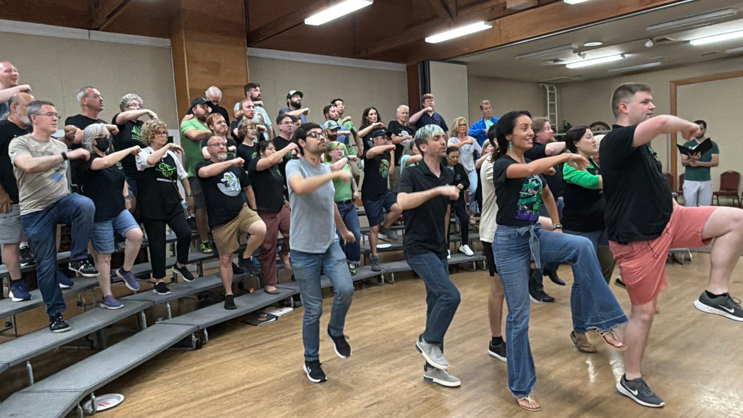 Group of people participating in a dance or exercise class in a room with wooden floors and tiered seating.