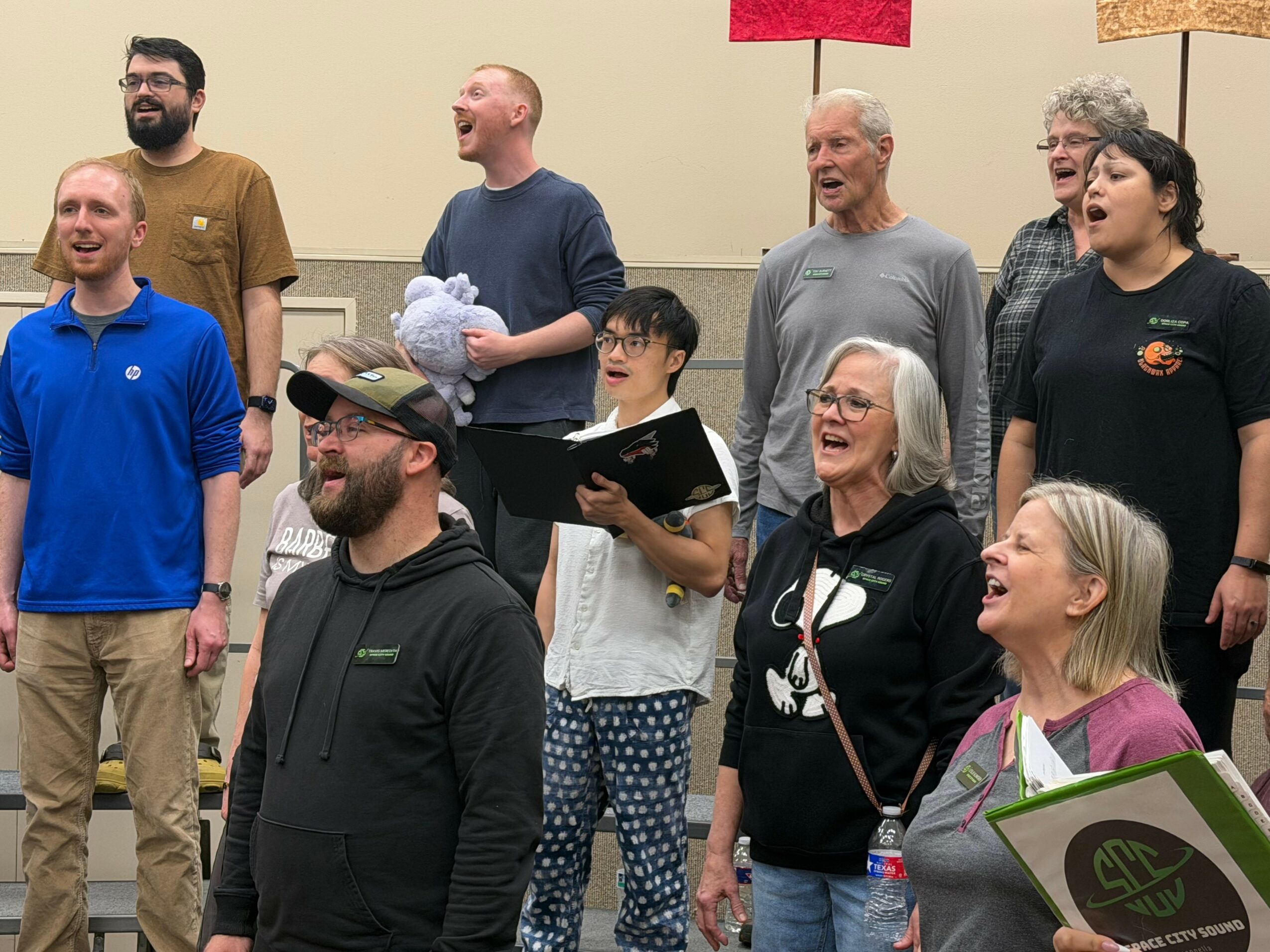Group of nine people singing together, some holding music sheets, in a room with beige walls.