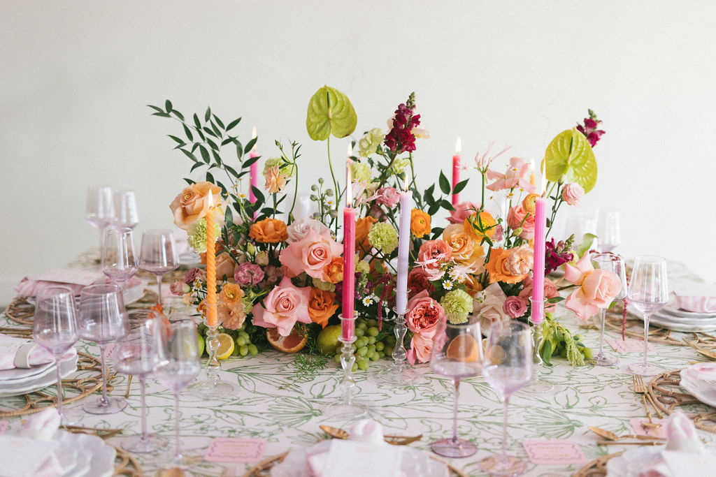 Elegant table setting with a floral centerpiece, pink and orange flowers, candles, and glassware, on a white tablecloth.