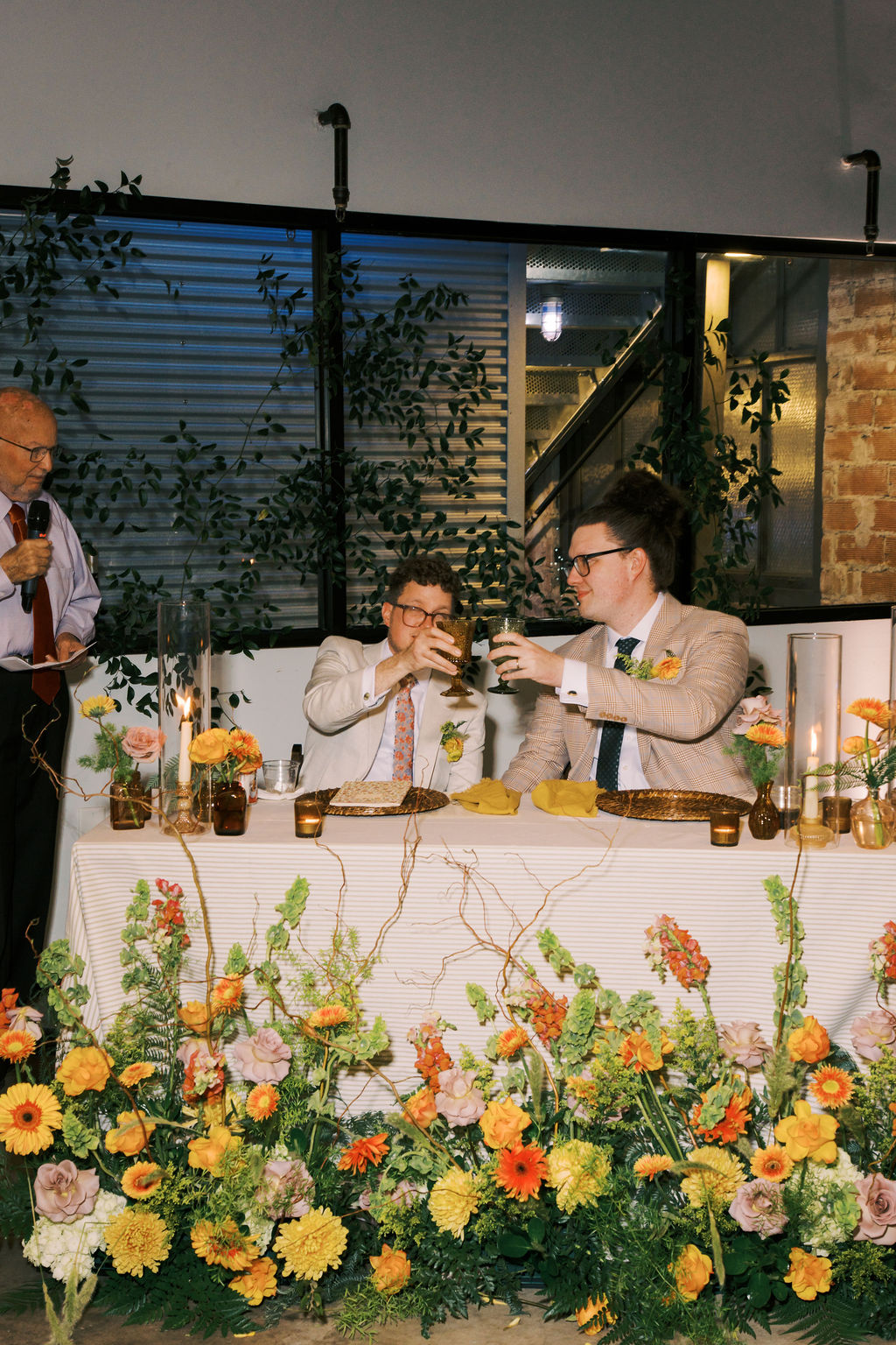 Two men sit at a decorated table with flowers, raising glasses in a toast, with a person standing nearby indoors.