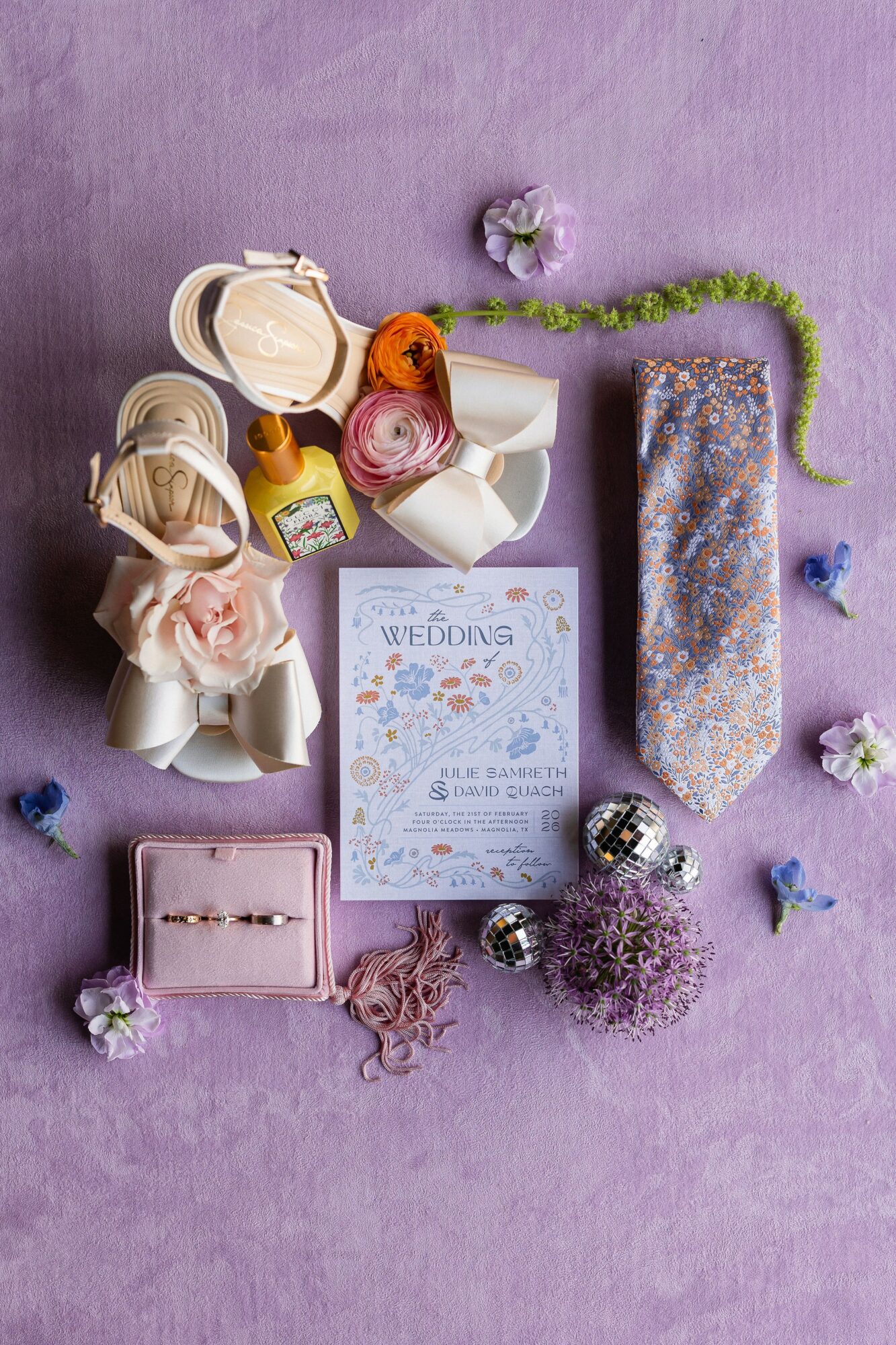 Wedding accessories including rings, flowers, jewelry, and a gift box arranged on a purple surface.