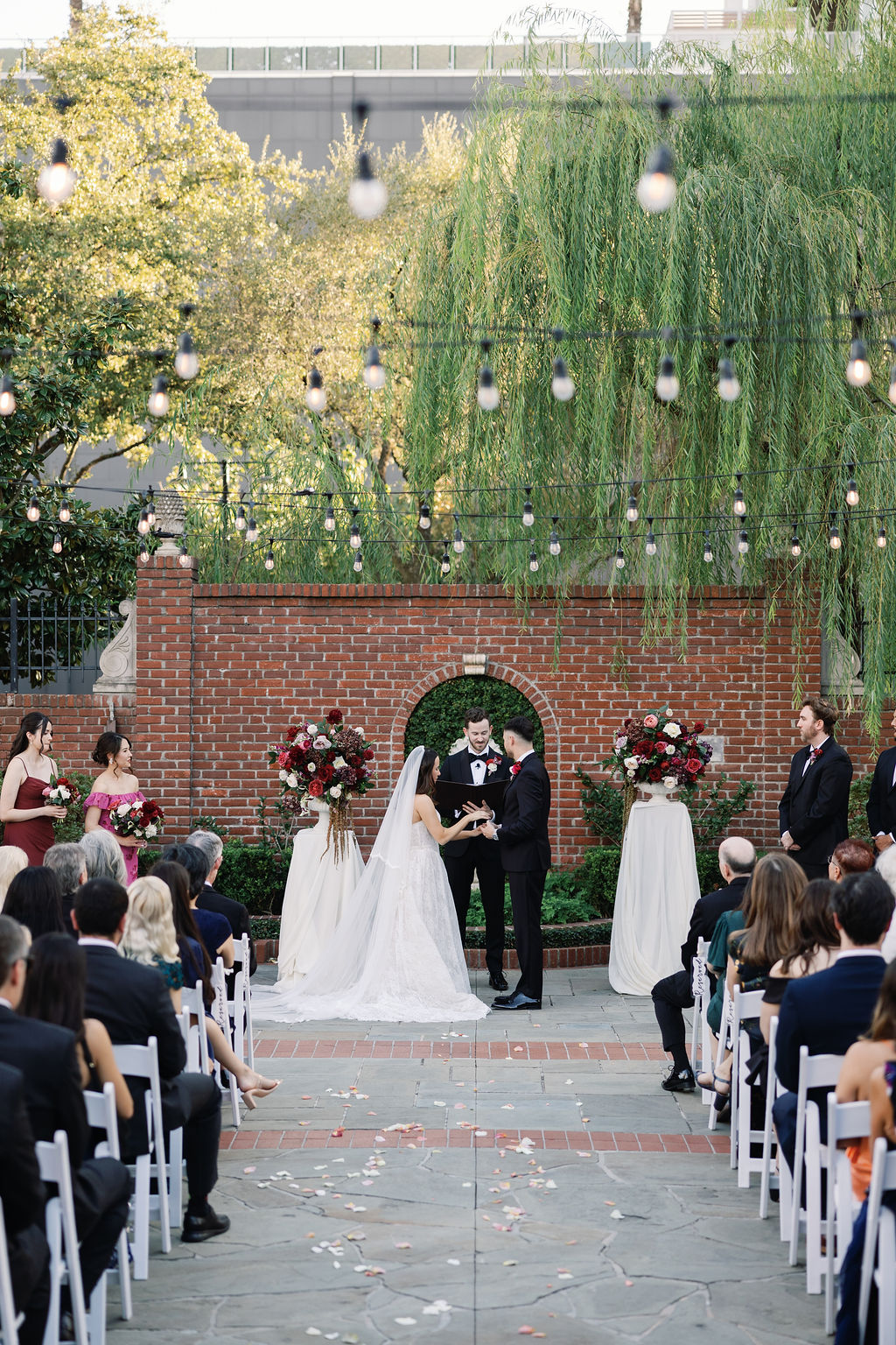 Outdoor wedding ceremony with bride and groom standing before guests, decorated with flowers and string lights, brick wall background.