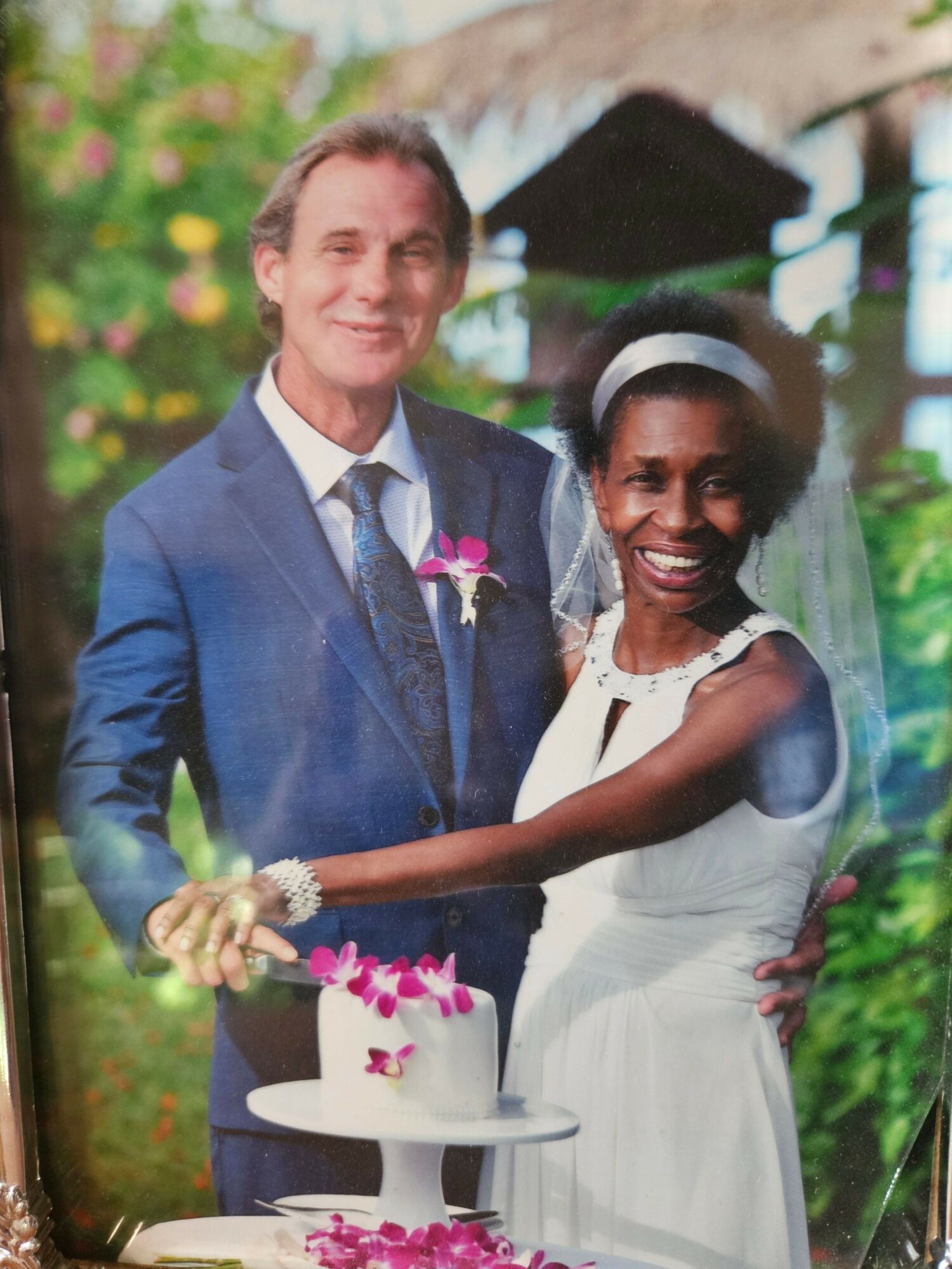 Two women, one in a white dress and the other in a suit, cutting a wedding cake outdoors with greenery in background.